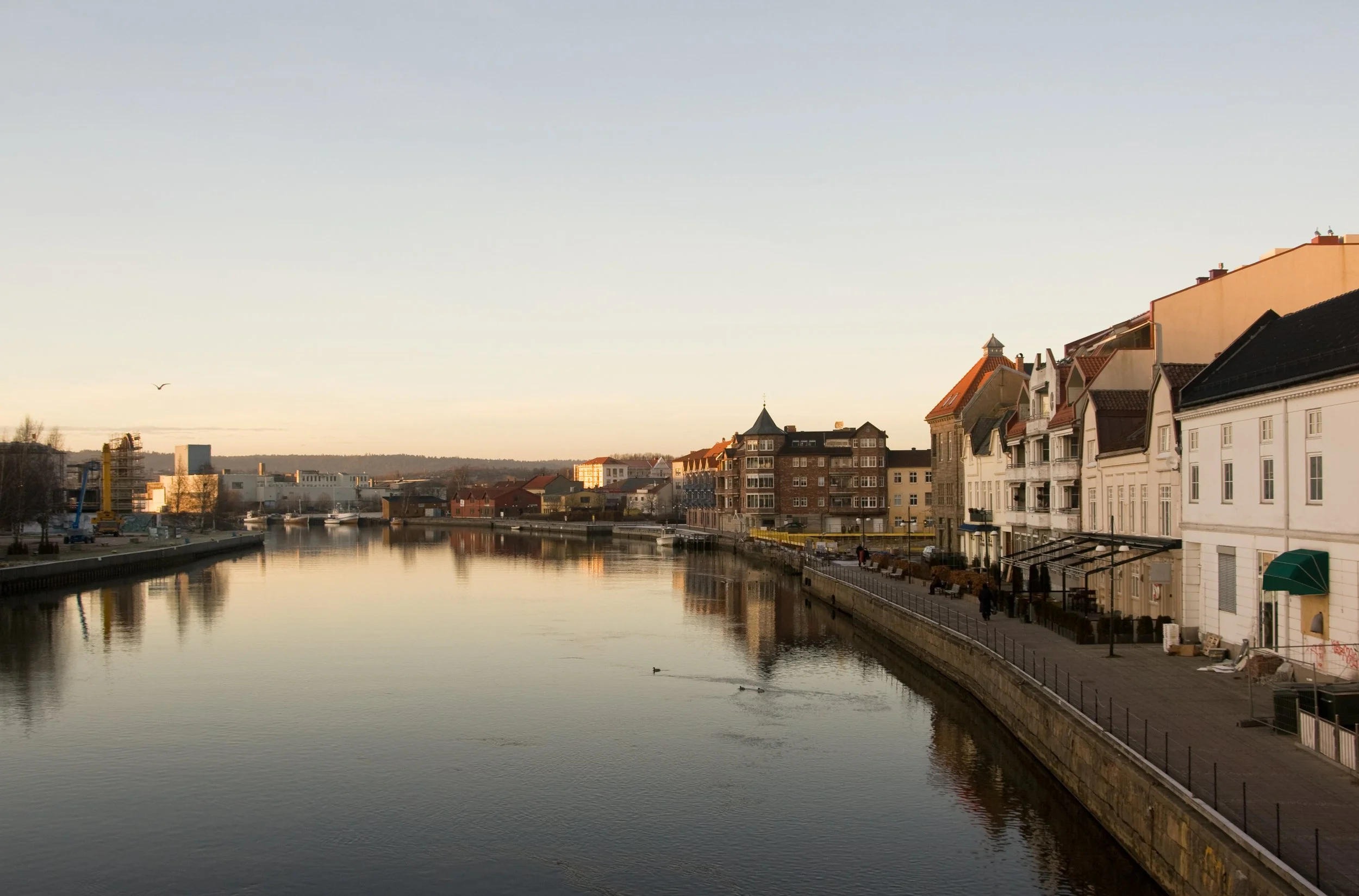 Bygninger langs en kanal i Fredrikstrad i solnedgang, med en rolig vannflate og noen mennesker på fortauet.
