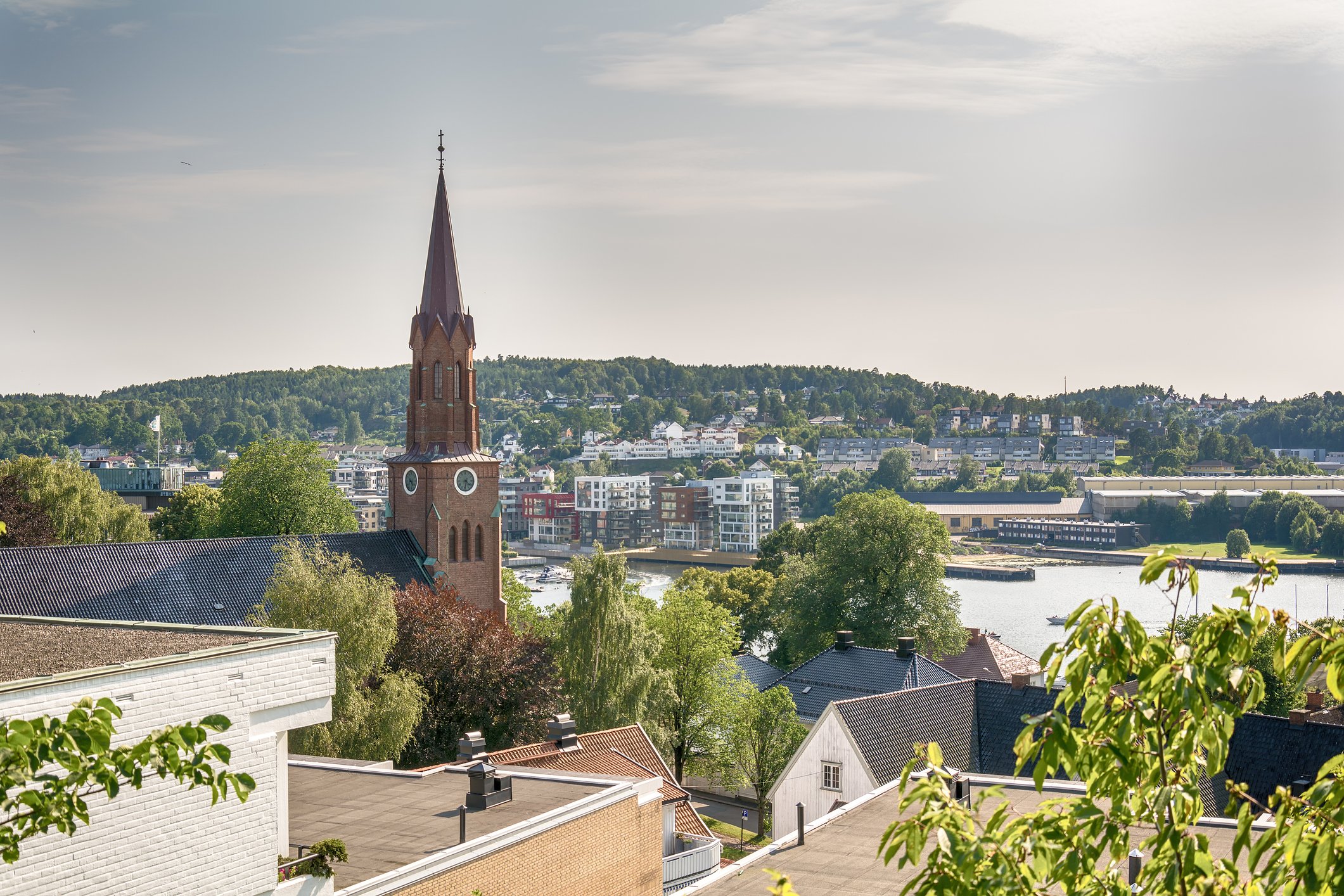 Utsikt over en by med en rød kirketårn i forgrunnen og moderne bygninger i bakgrunnen, omgitt av grøntområder ved en elv.