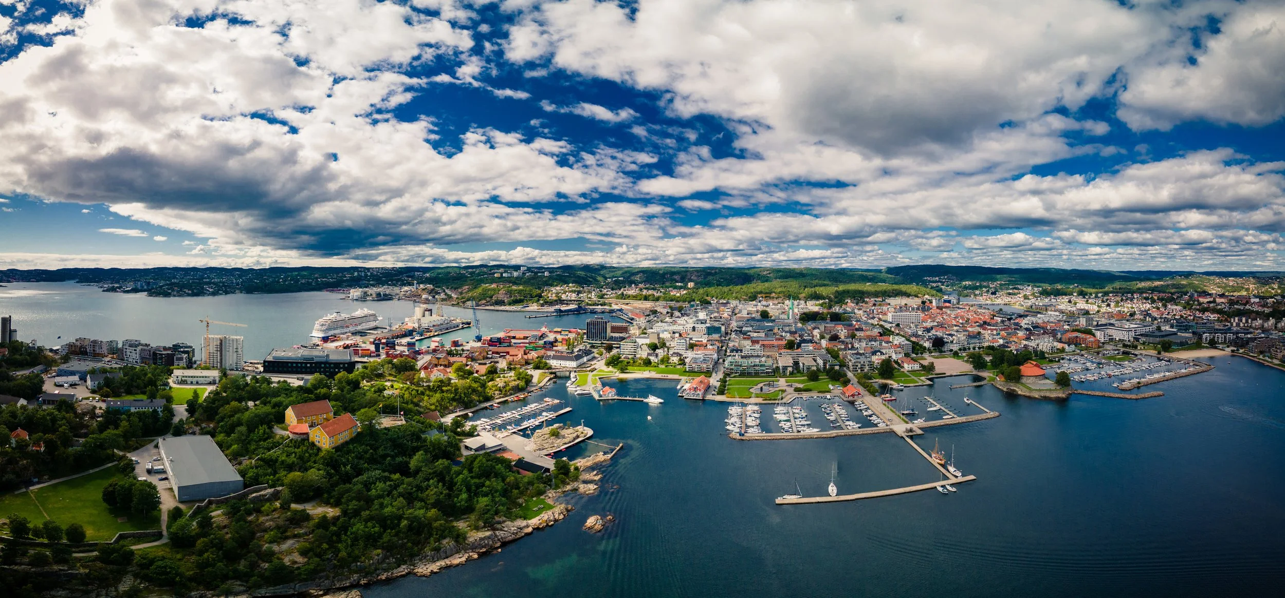 Oversiktsbilde av en havneby med båter og kraner, omgitt av grønne områder og bygninger, under en delvis overskyet himmel.