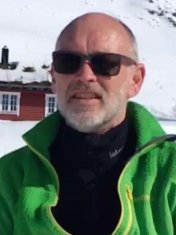 En mann med solbriller og grønn jakke som står foran snødekt fjelllandskap.