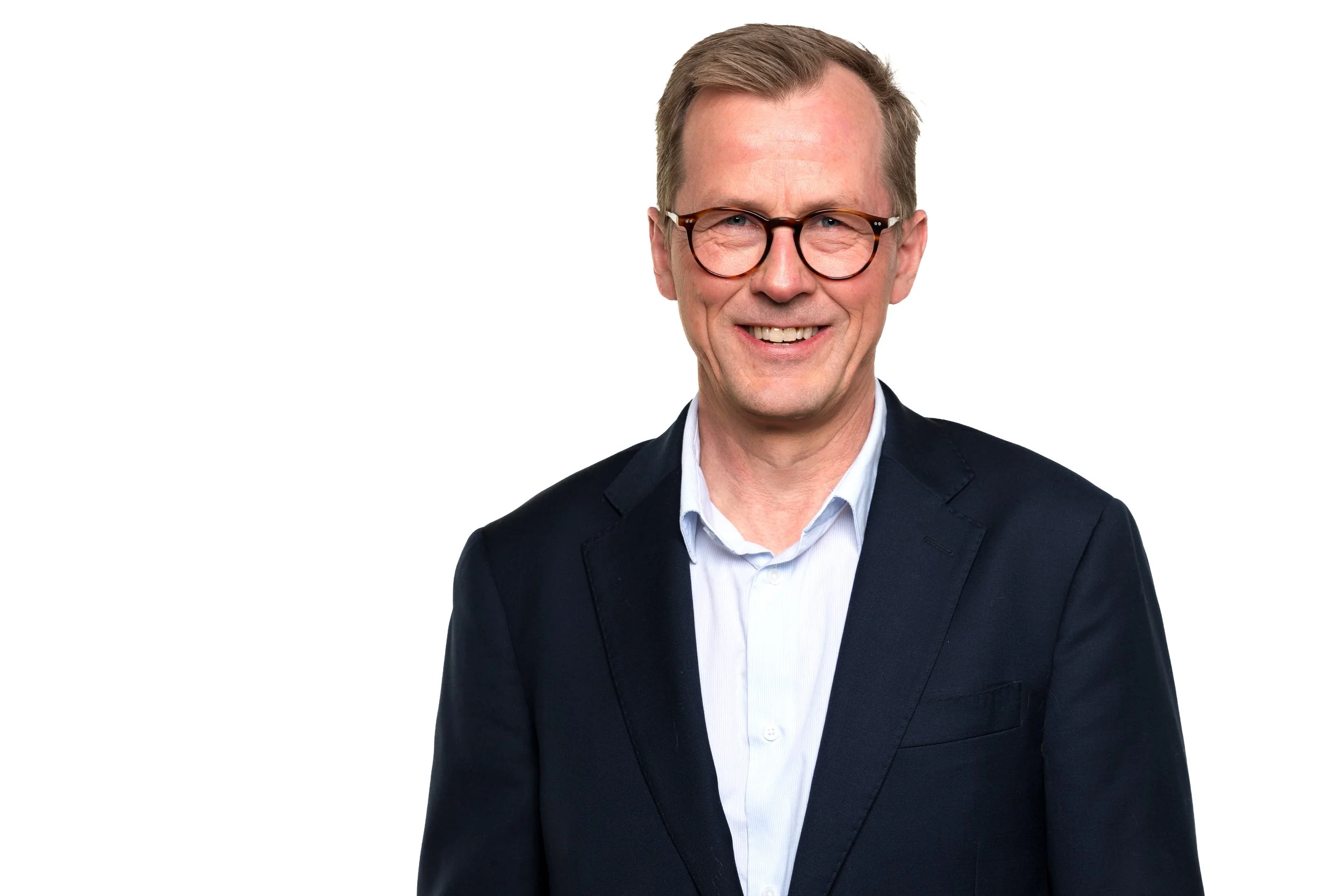 Portrett av en mann med brunt hår, briller og smilende, iført en mørk dress og hvit skjorte, mot hvit bakgrunn.