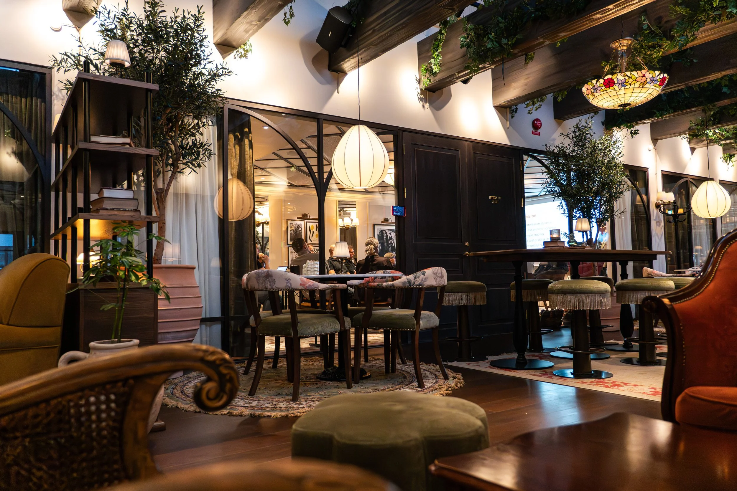 Innendørs restaurant med bord, stoler, store lyslykter og grønne planter, med mennesker som sitter og snakker.