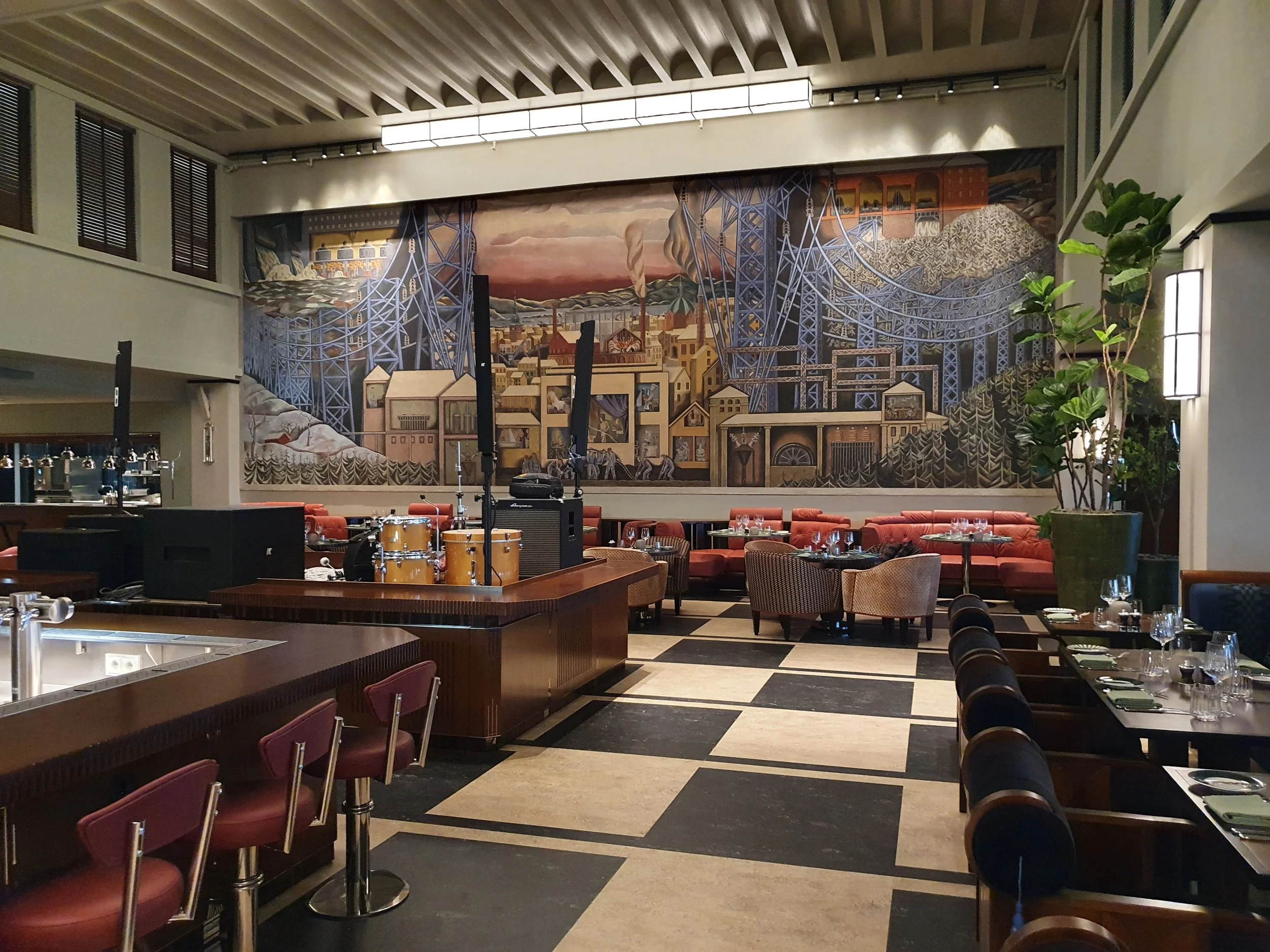 Interiør av en restaurant eller kafé med bord dekket for servering, blomsterpotter og en stor muralmaleri på veggen av en industribanelinje med en bakgrunn av fjell og flere bygninger.