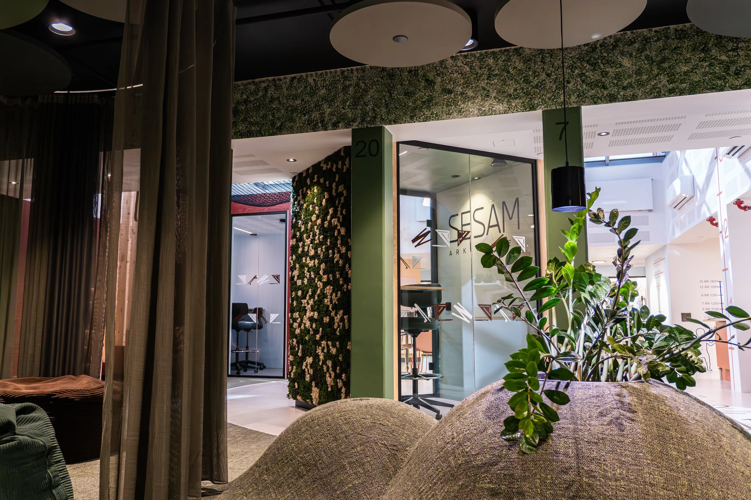 Innendørs område med grønne planter, glassvegg med tekst 'SESAM Arkitektur', samt gulv og vegger i lys farger, moderne møbler og dekorative elementer.