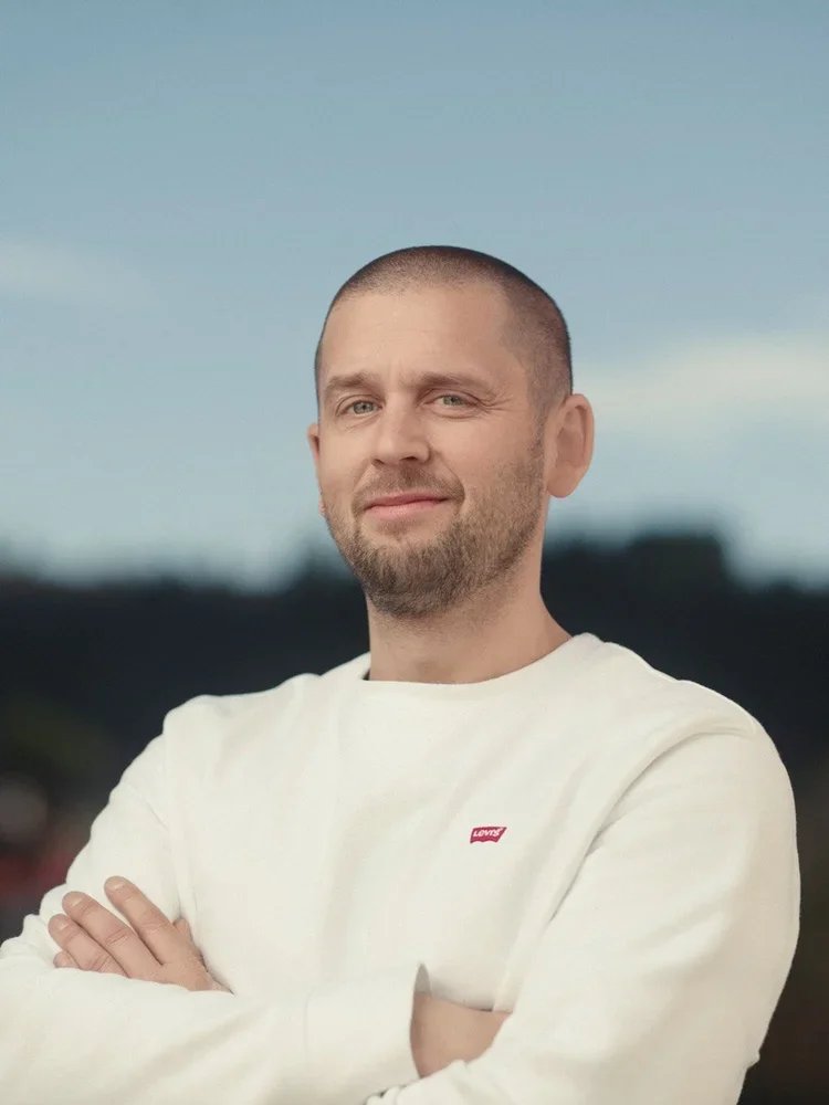 En mann med kort hår og skjegg som står med armene krysset, iført en hvit genser med Levi's-logo, utendørs under en klar himmel.