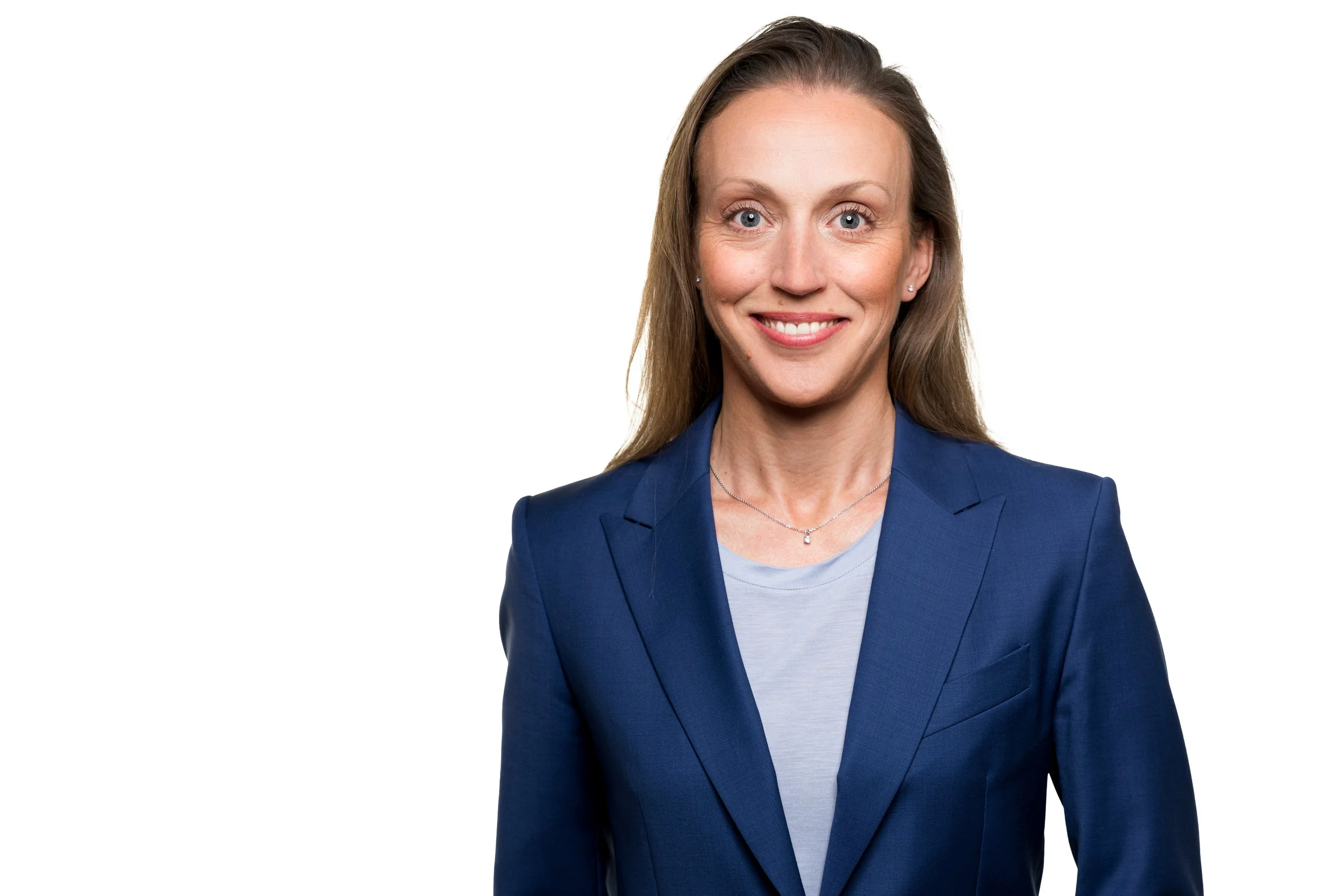 Portrett av en smilende kvinne med langt mørkt hår, iført blå blazer og lyse T-skjorte, mot hvit bakgrunn.