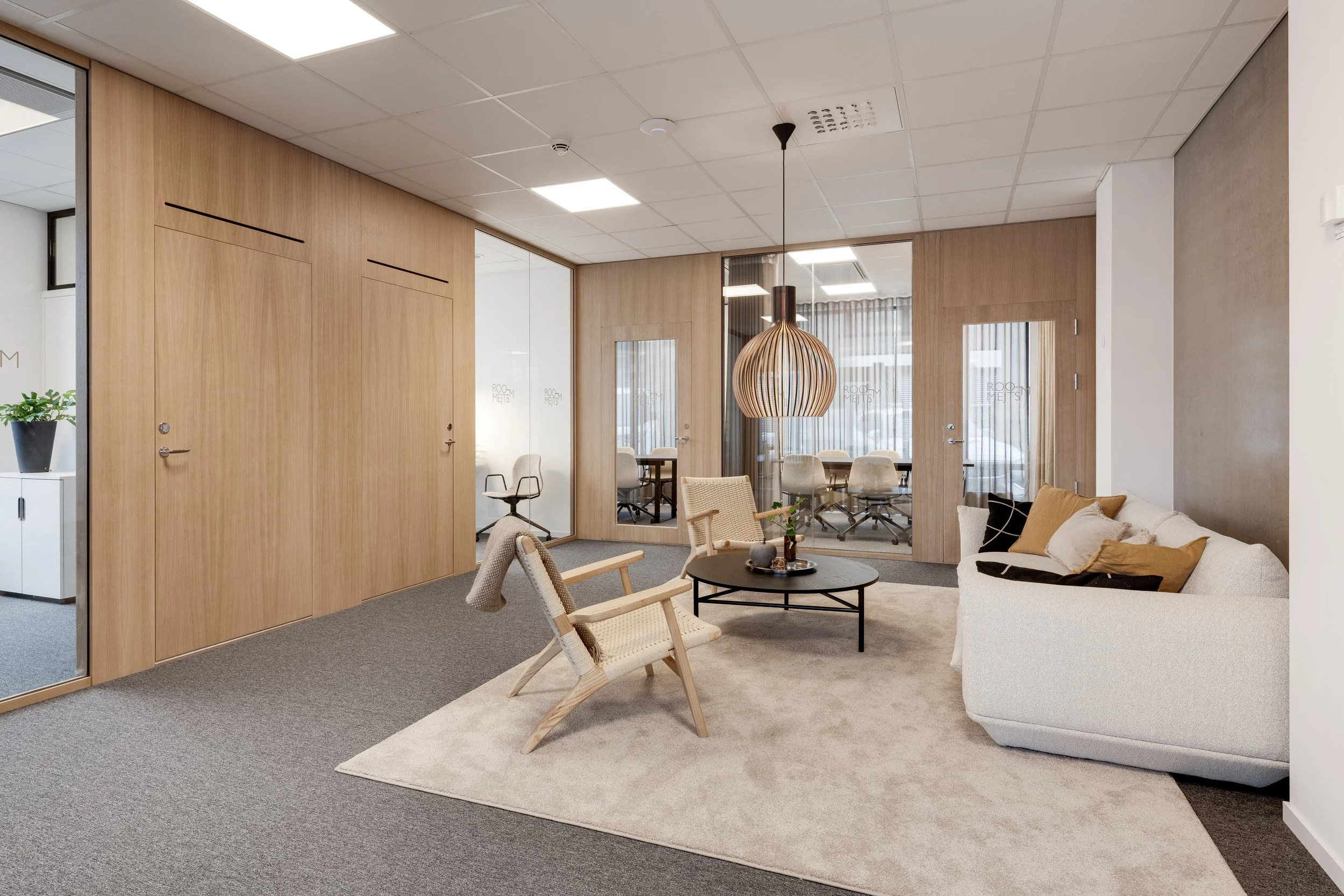 Et moderne kontor- eller venterom med komfortable stoler, sofa, lavt bord, store vinduer med gardiner og en sentral takpendel i tredesign.