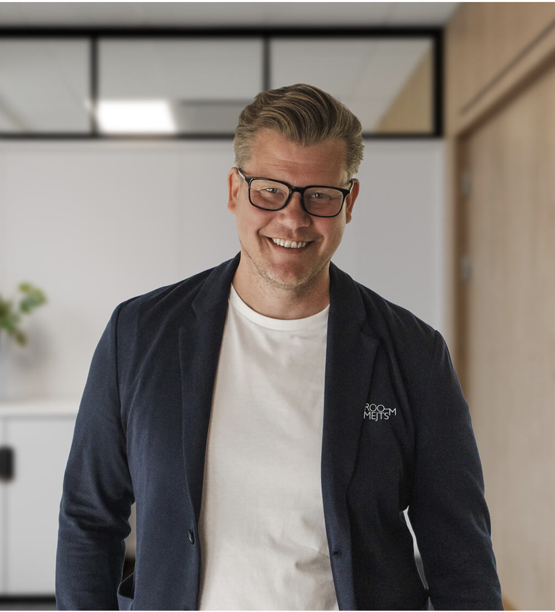 En mann smiler, har på seg briller, hvit t-skjorte og mørk blazer, står innendørs i et rom med speil og trepanel på veggen.