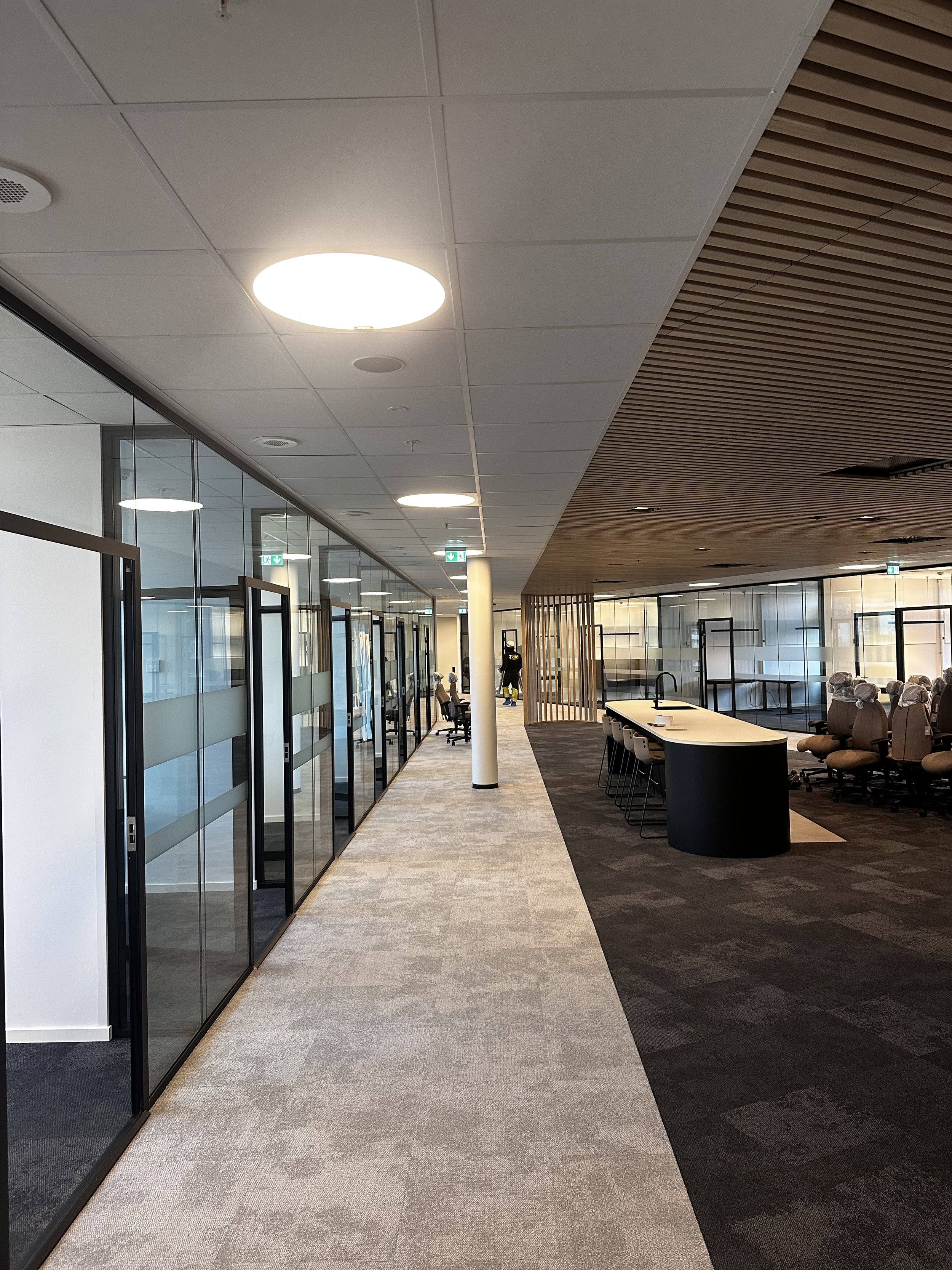 Moderne kontor- eller resehall med glassdører, sittegrupper, barstoler og lamper, med tre personer i bakgrunnen.