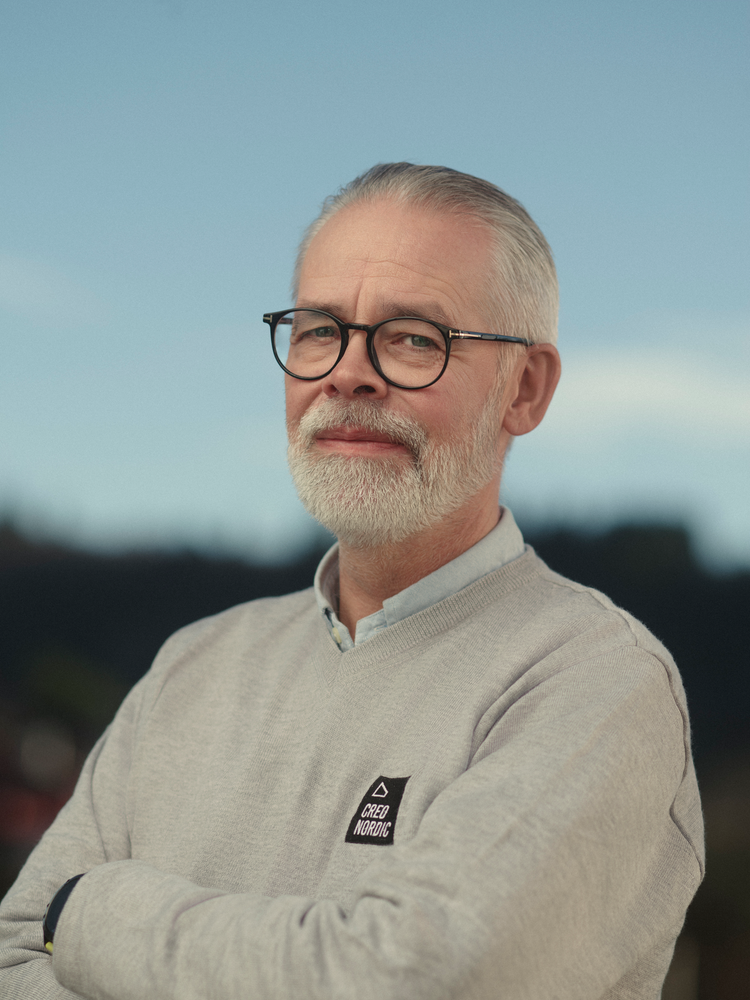 En eldre mann med grått hår og skjegg, iført briller og en lys grå genser med 'Cred Nordic'-logo, står ute mot en mørk bakgrunn med en blå himmel.