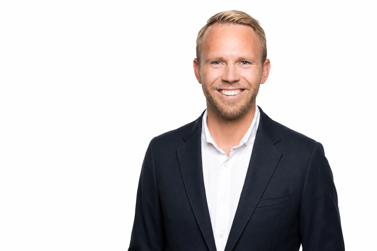 En mann med lyst, kort hår og skjegg, iført dress og hvit skjorte, smiler mot kameraet. Flat hvit bakgrunn.
