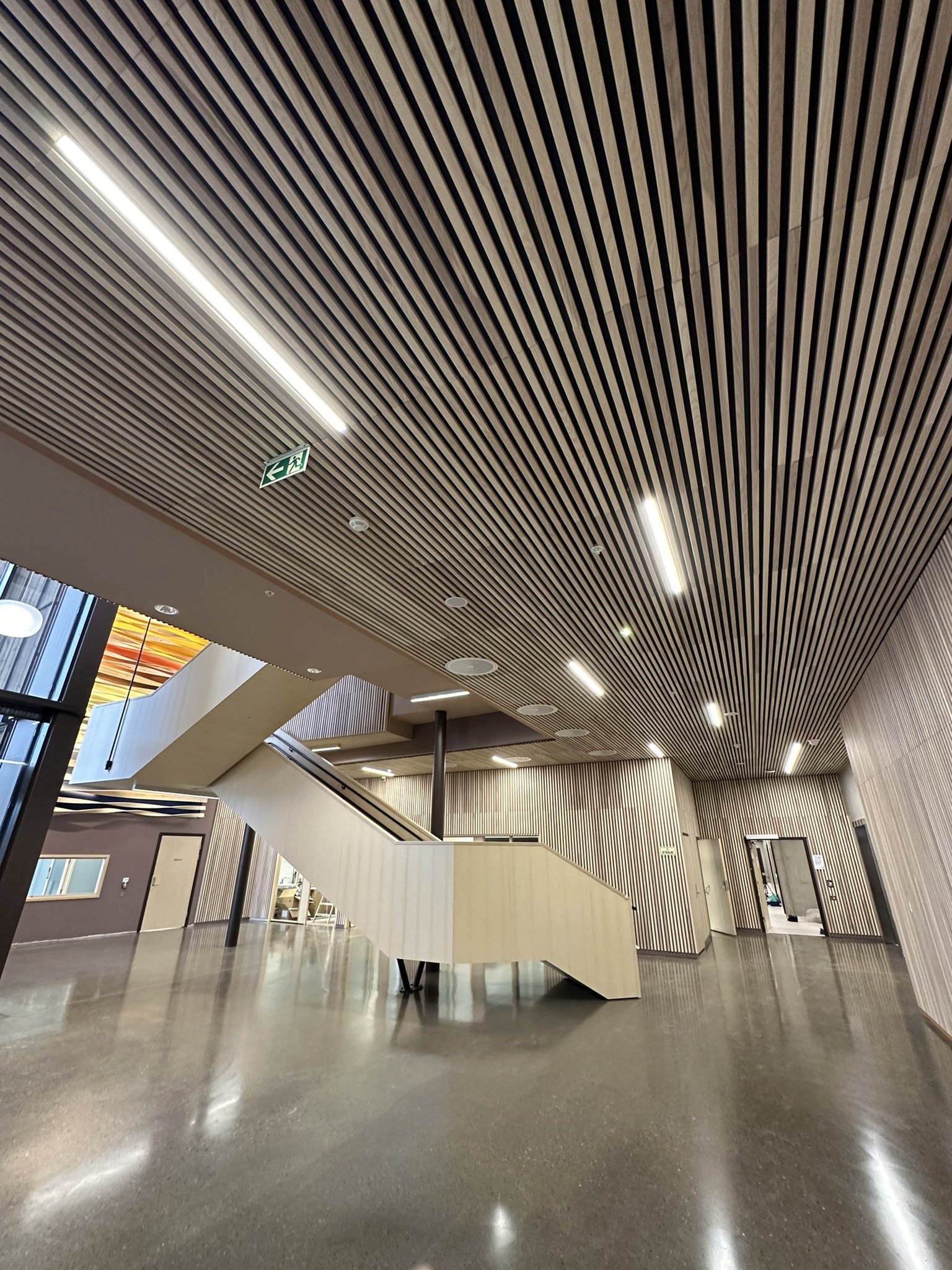 Innendørs med moderne tak med parallelle trepaneler og innebygde lys, trapp med glassrekkverk og reoler langs veggene.