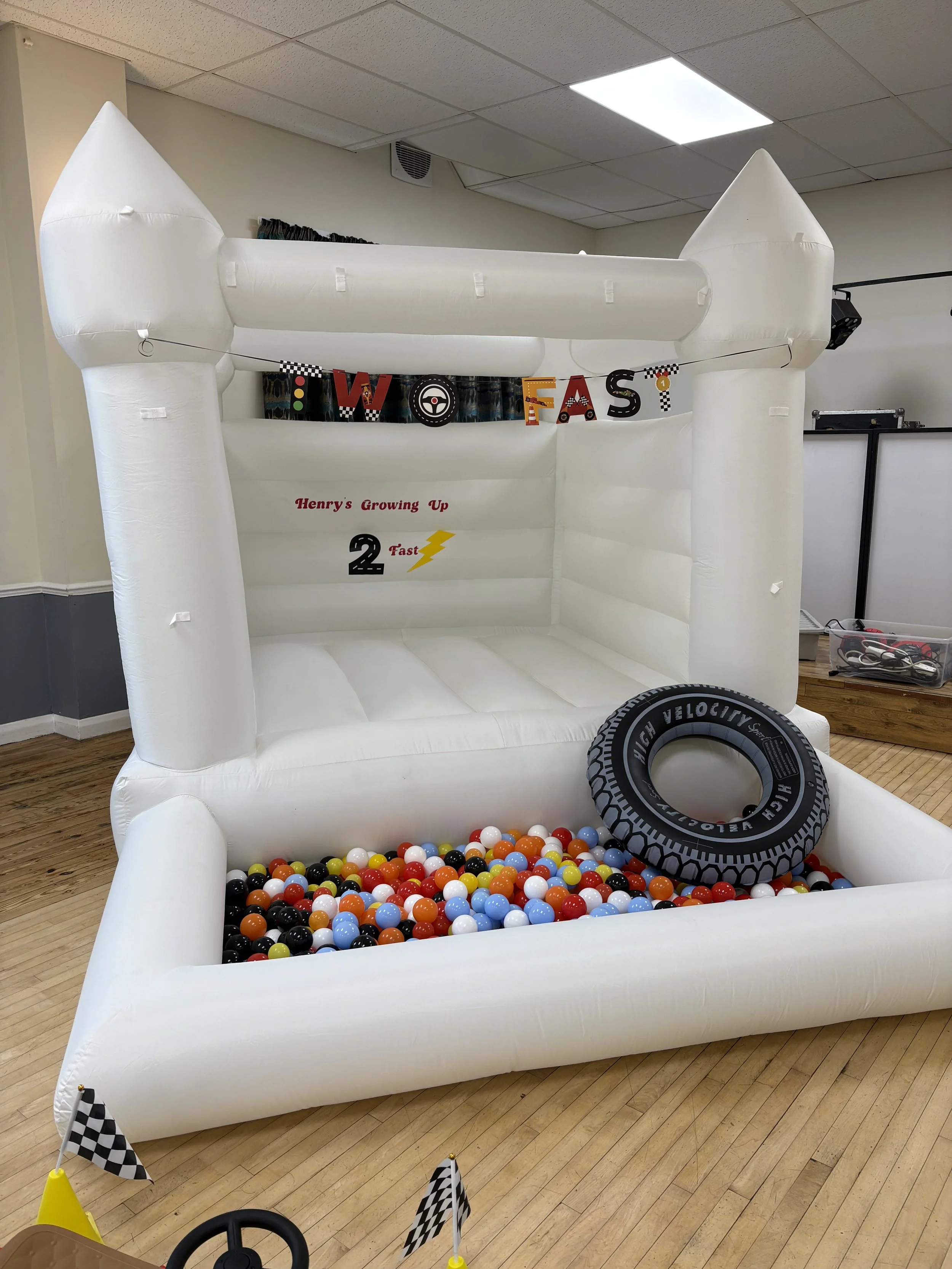 An inflatable white castle-style bounce house with a ball pit filled with colorful plastic balls, a black rubber tire, and race flags as decoration, set indoors on a wooden floor.