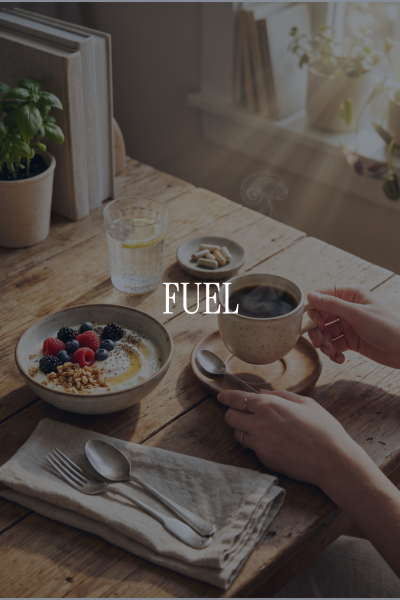 A cozy breakfast scene with a cup of coffee, a bowl of yogurt topped with berries and granola, a glass of water with lemon, a small plate with pills, and a spoon and fork on a napkin on a wooden table near a window with plants.