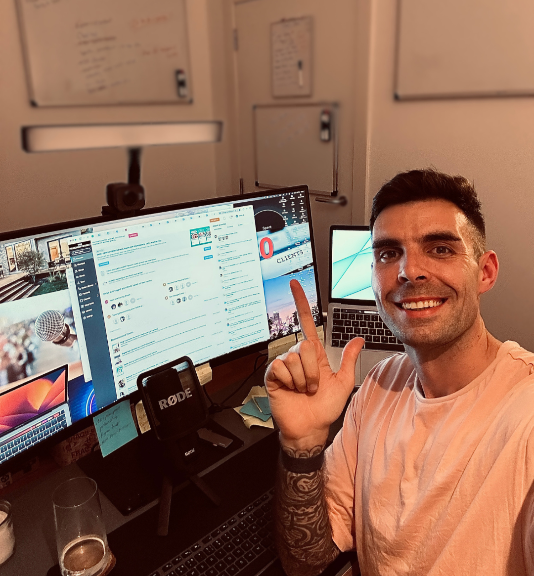 Man taking a selfie at a cluttered desk with two monitors, a MacBook, a microphone, and a RØDE speaker, pointing to the screen on the right, smiling, with whiteboards in the background.