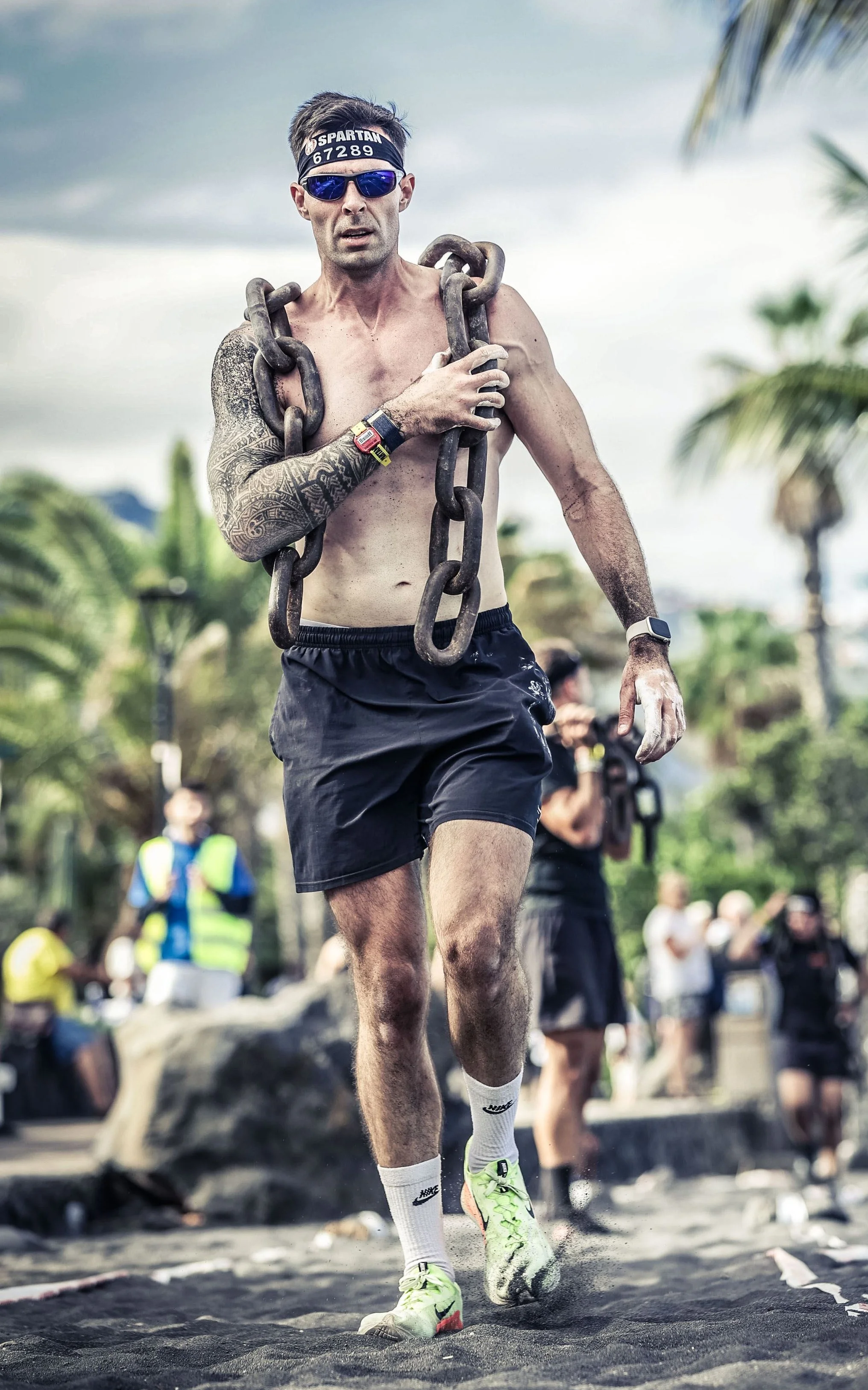 A shirtless male runner wearing sunglasses, a headband, black shorts, and bright yellow running shoes, running outdoors with spectators in the background.