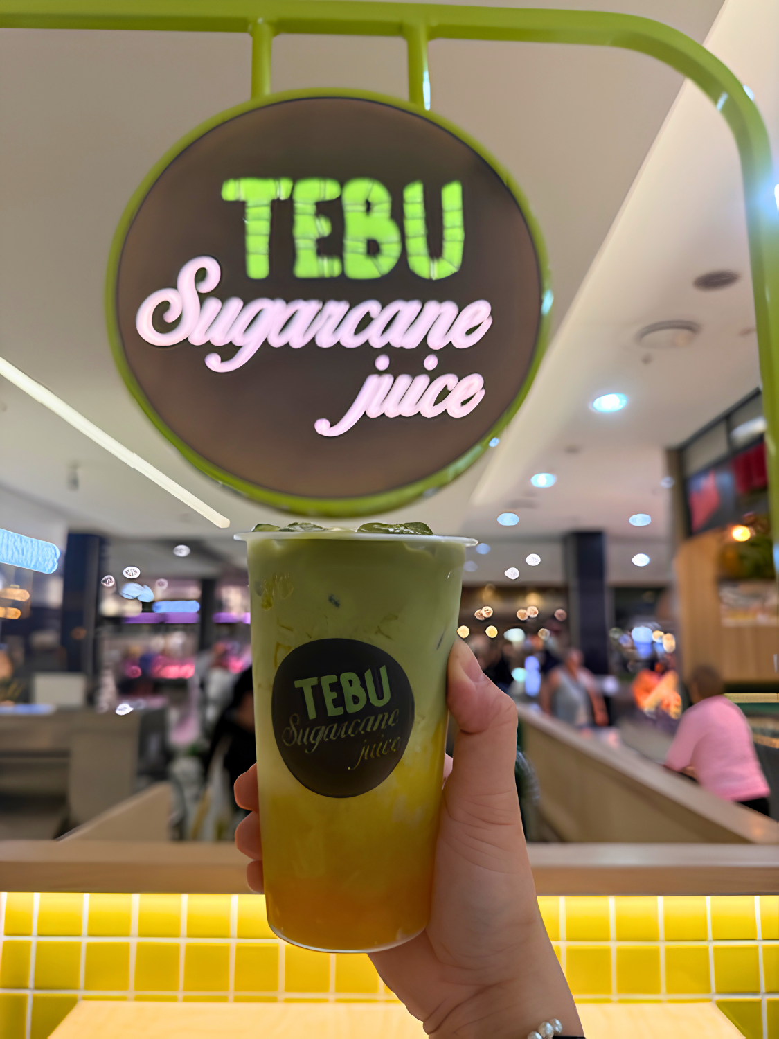 A hand holding a green colored juice in a clear cup with the label 'TEBU Sugarcane juice'. Behind is a bright, modern food court and a circular sign displaying the same brand name and product.
