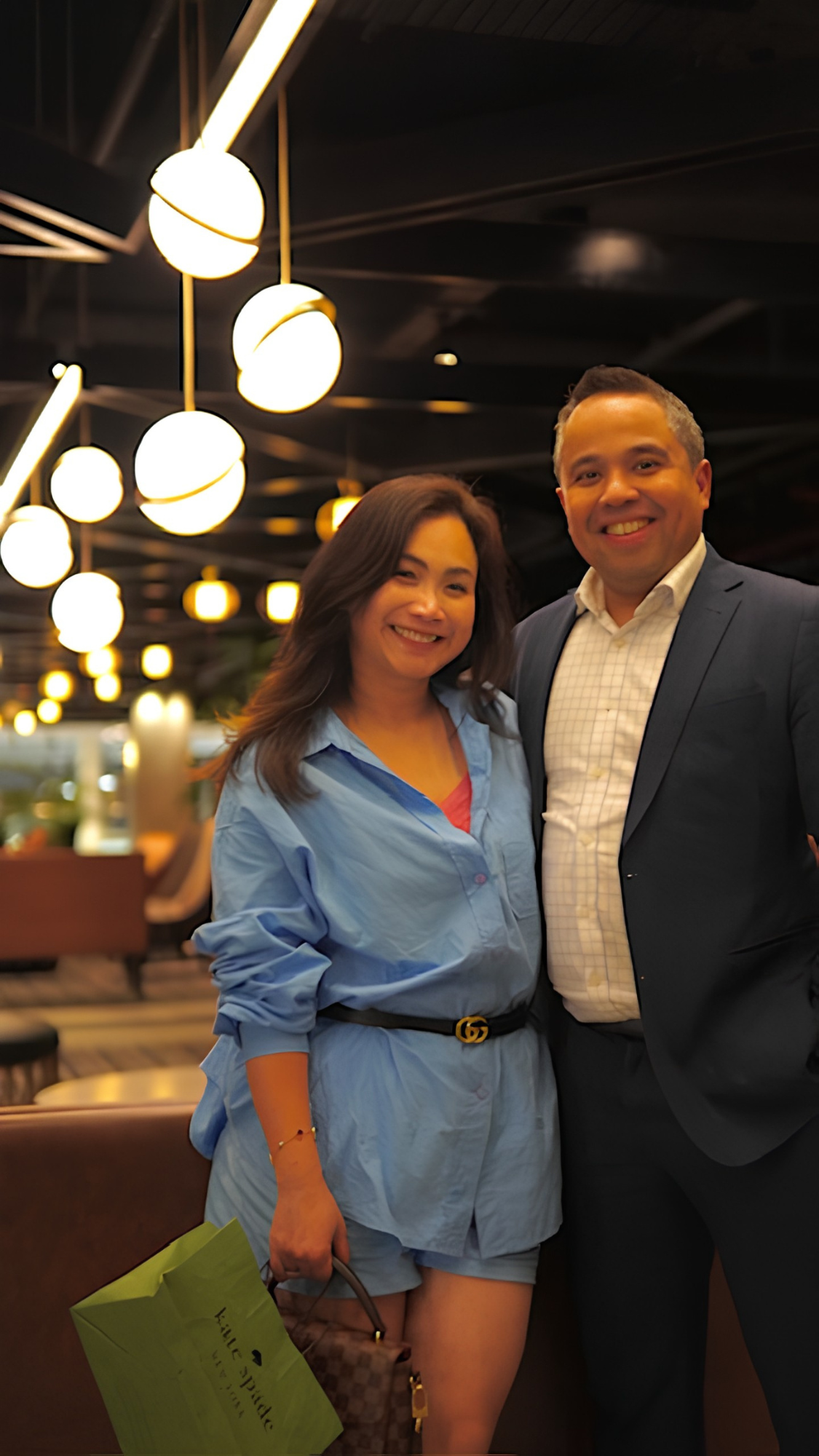 A smiling woman in a blue shirt and shorts standing next to a man in a dark suit inside a restaurant or lounge, with decorative lighting on the ceiling.