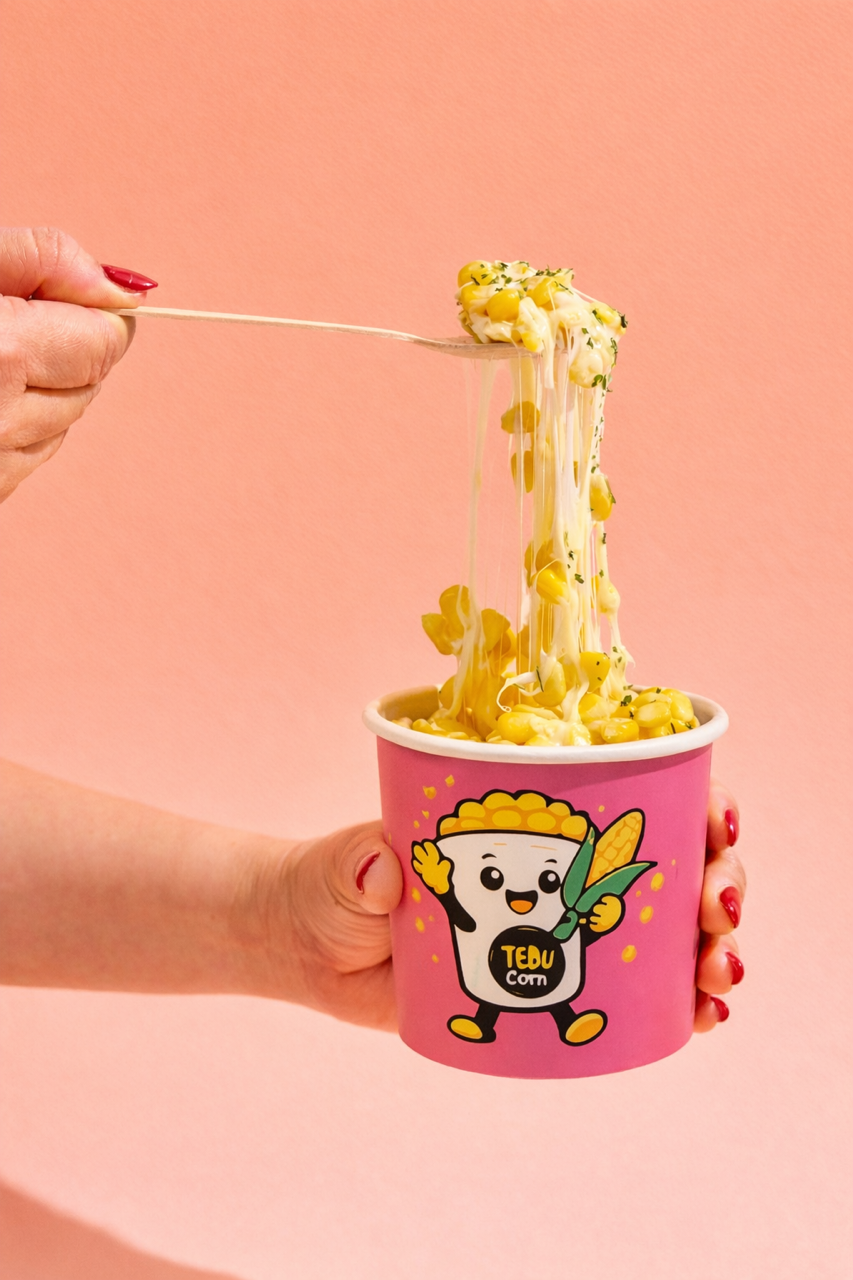 A person with red nail polish holding a pink cup with a cartoon character and the words "TEDU CORN" on it. The cup contains cheese popcorn with melted cheese stretching as a hand lifts a scoopful.