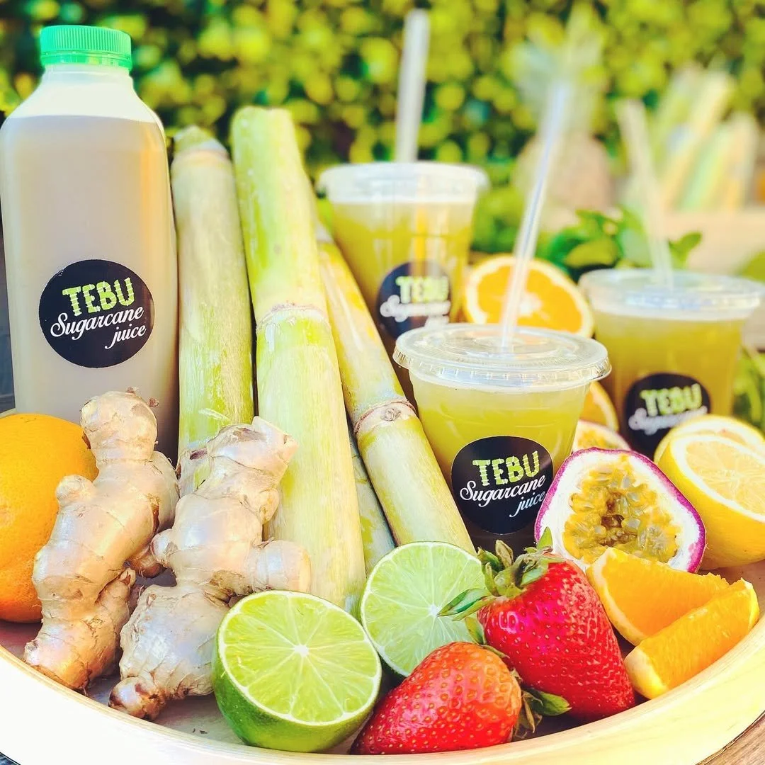 A variety of fresh fruits and vegetables, including ginger, lemons, limes, strawberries, passion fruit, and oranges, along with bottles of sugarcane juice and cups of juice from TEBU Sugarcane Juice, arranged outdoors.