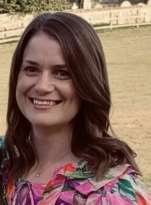 A smiling woman with long brown hair wearing a colorful floral top outdoors.