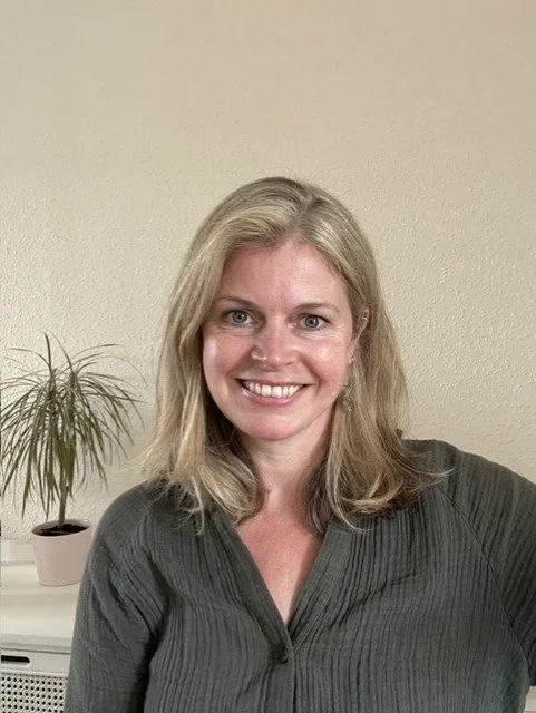 A smiling woman with blonde hair wearing a dark green shirt, sitting in front of a beige wall with a plant on a desk behind her.