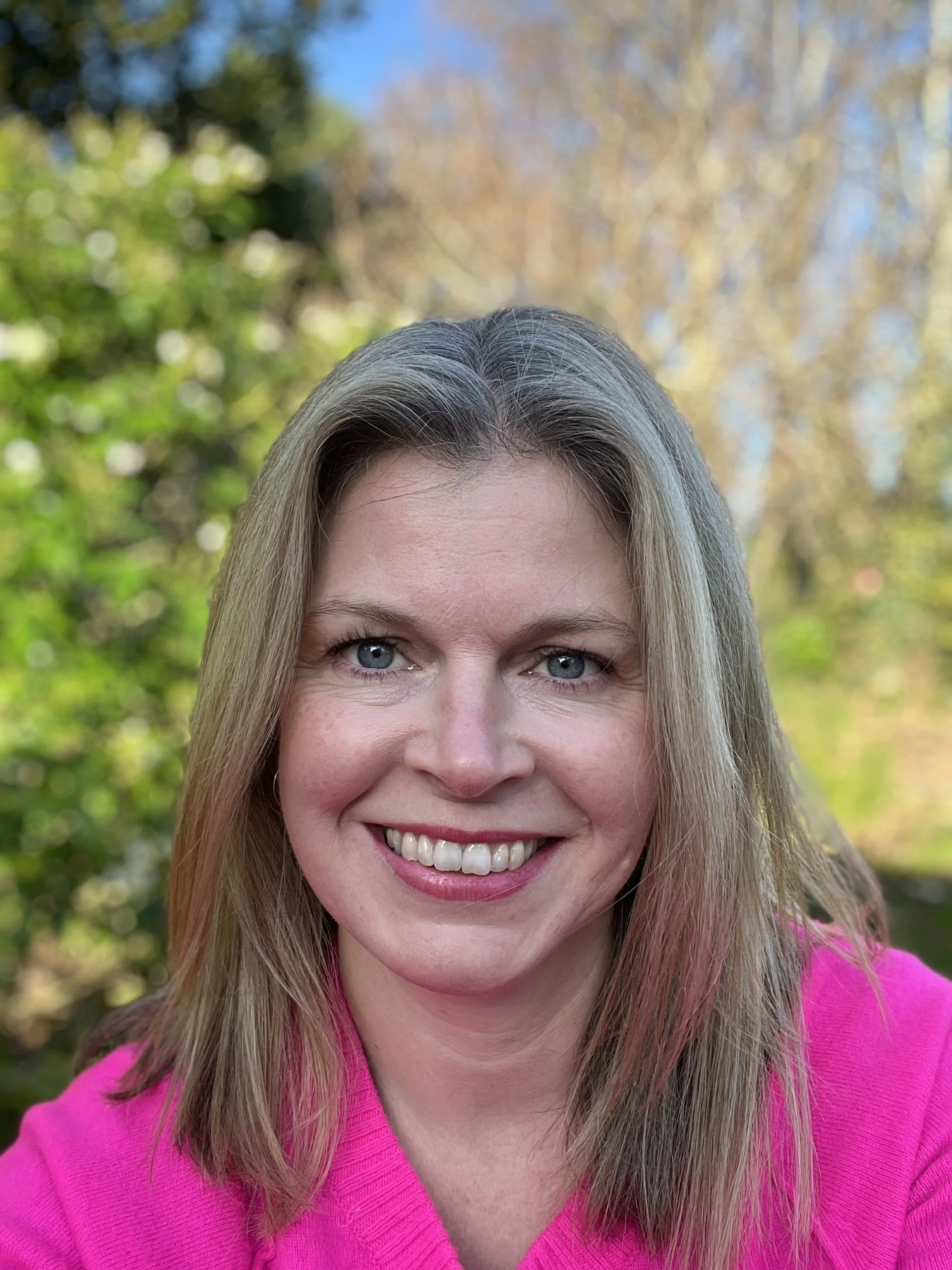 Close-up of a smiling woman with light blonde hair and blue eyes, wearing a pink top, outdoors with blurred green and brown trees in the background.