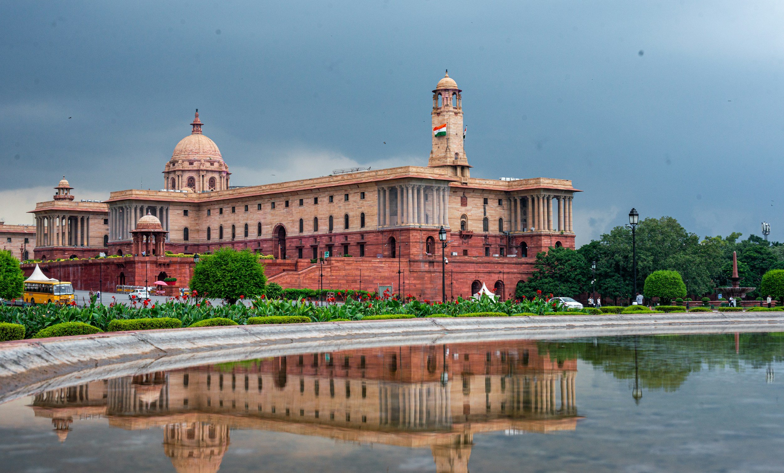 Historical government building with domes and a flag, surrounded by greenery and street activity, with a cloudy sky overhead. Reflection in a water body in the foreground.