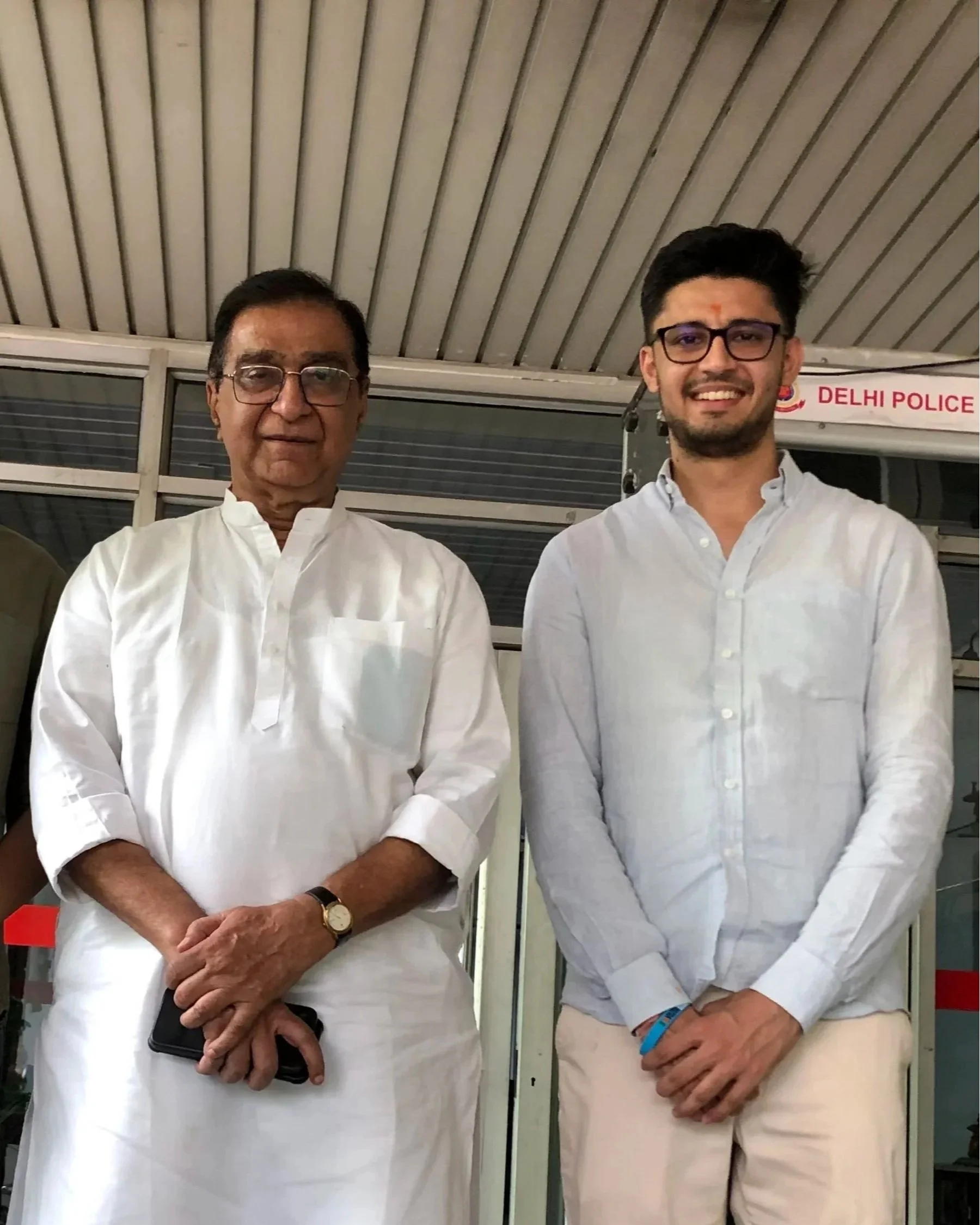 Two men standing side by side in front of a wall with a sign that reads 'Delhi Police.' Both are dressed in white clothing and are wearing glasses. They are smiling, with one of them holding a phone.