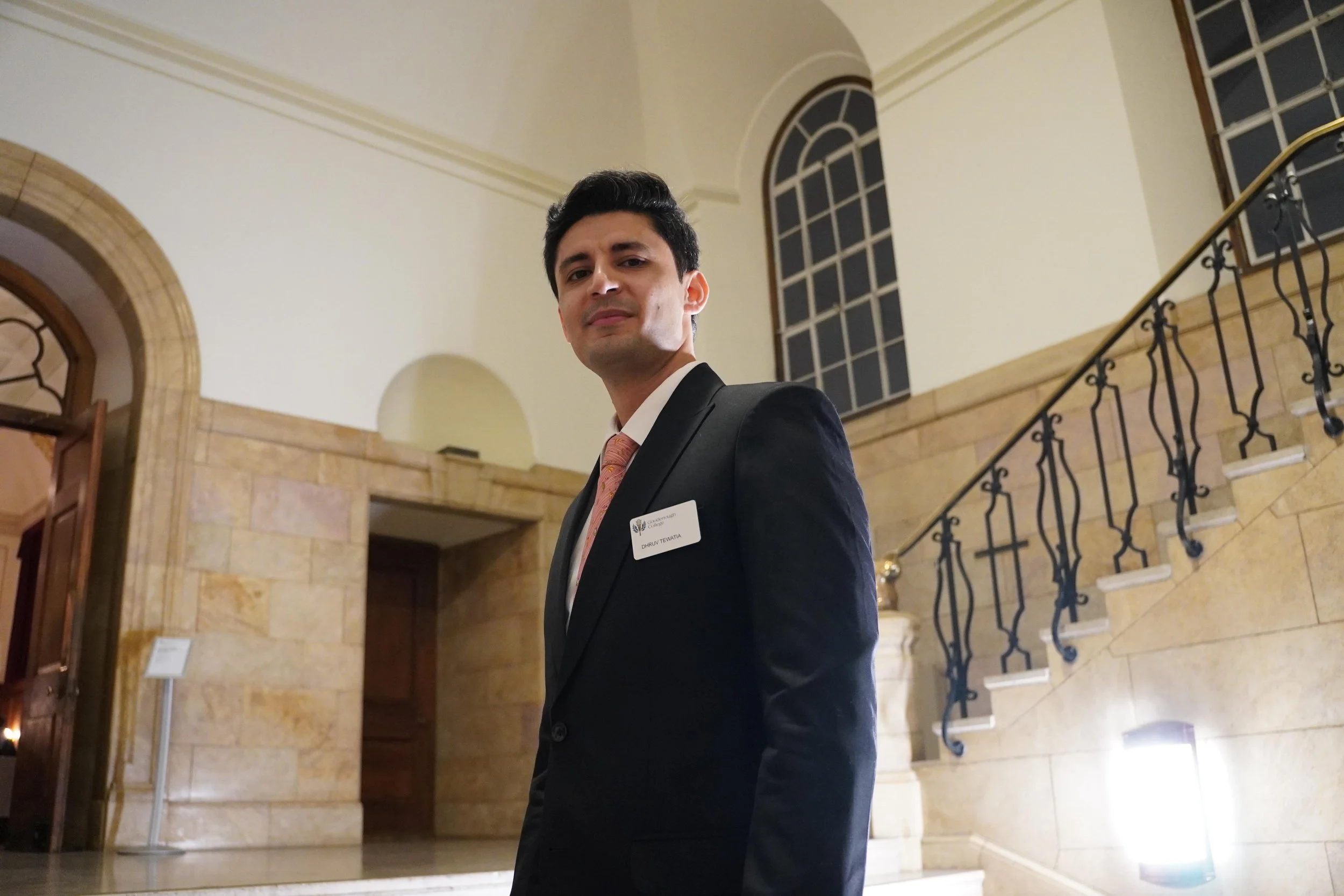 A man in a black suit and pink tie standing inside a grand building with tall arched windows and a staircase with ornate black railing, marble walls, and warm lighting.