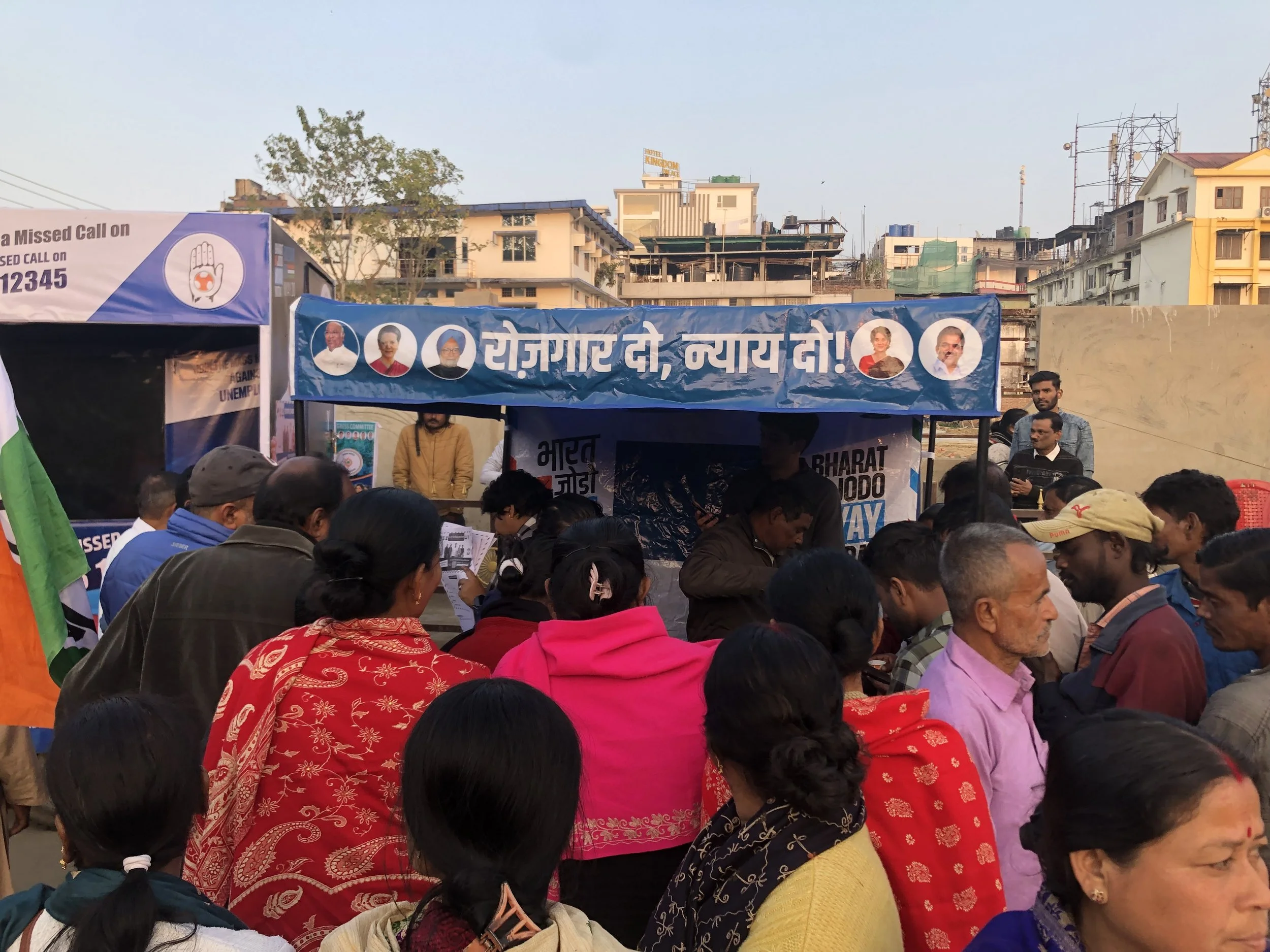 Crowd of people gathered outdoors with banners displaying political messages and images of political leaders in an urban setting.