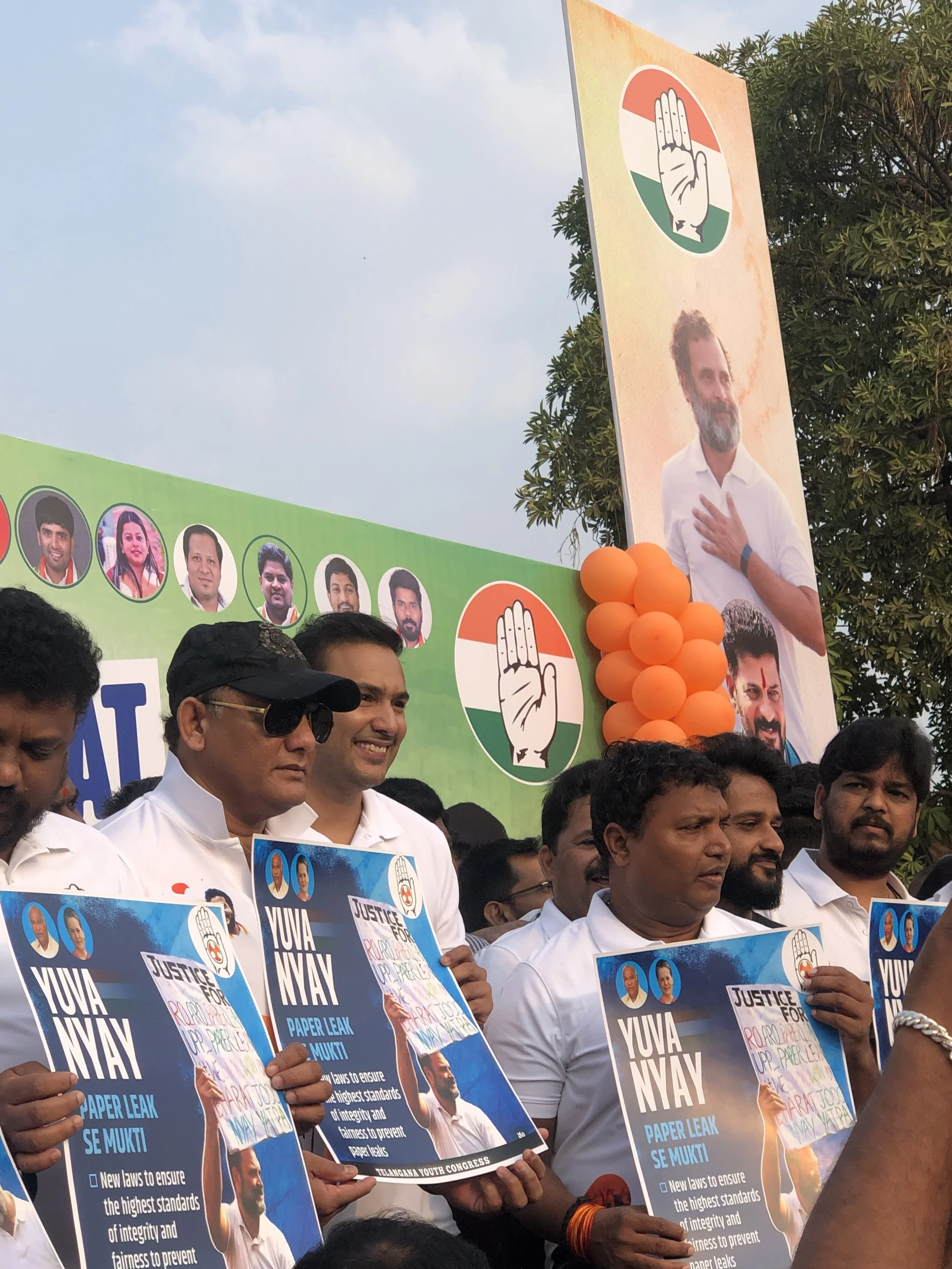 A group of people holding protest signs advocating for justice, integrity, and anti-corruption, with a large poster of a man on a stage behind them, featuring the Indian political party symbol of a hand, with orange balloons decorating the stage, indicating a political rally or demonstration.