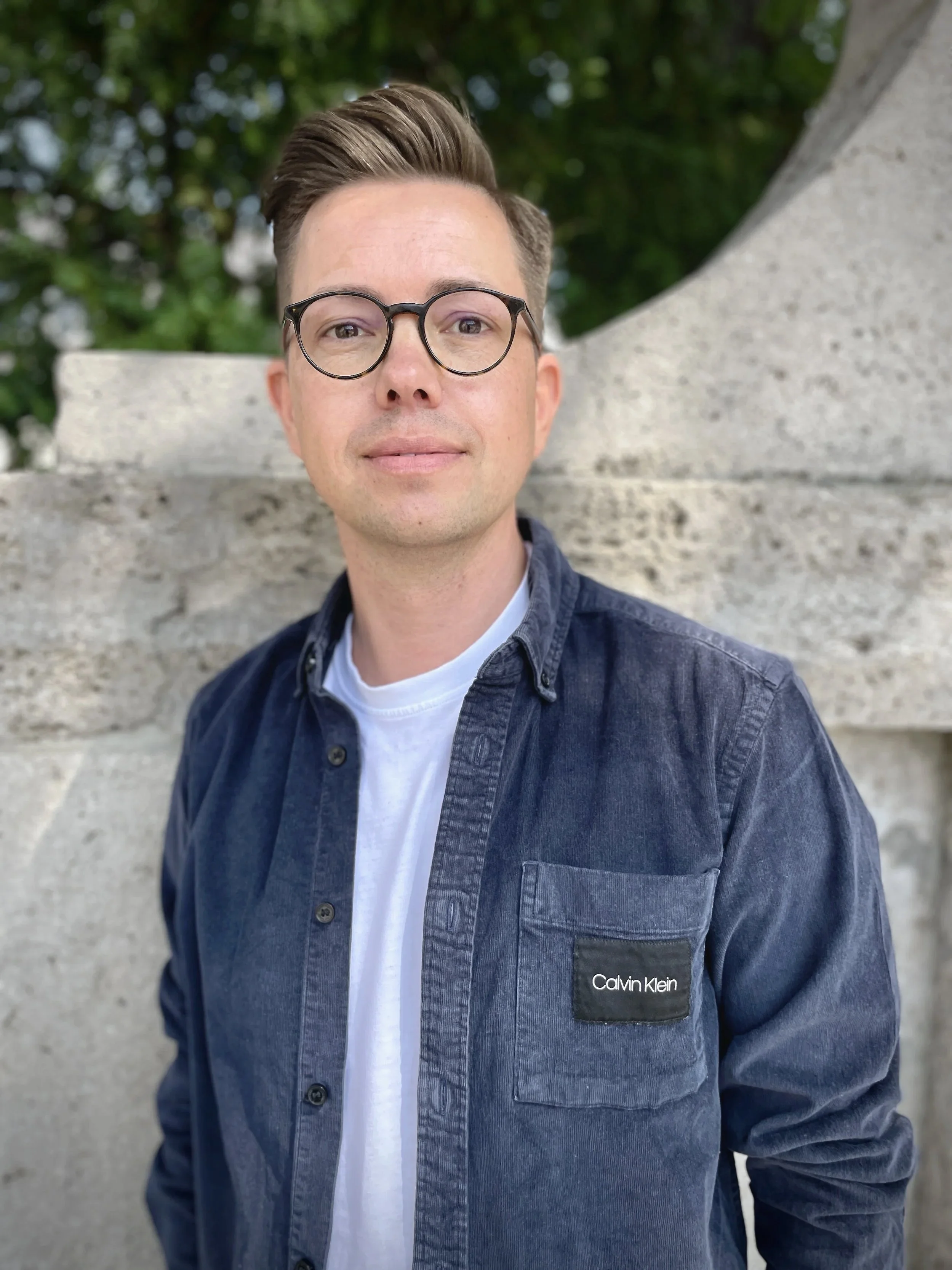 Nahaufnahme eines Mannes mit brünettem, gestyltem Haar und runden Brillen, trägt eine dunkelblaue Jeansjacke mit einem Calvin Klein-Logo auf der linken Brusttasche, steht vor einem grauen Steinwand, im Hintergrund grüne Blätter.