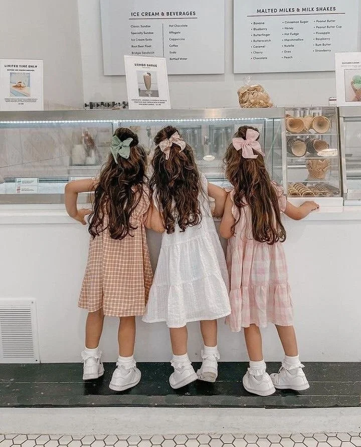 A charming vintage-inspired ice cream shop scene featuring three young girls in soft pastel summer dresses with oversized ribbon bows and classic white shoes standing heirloom-inspired clothing