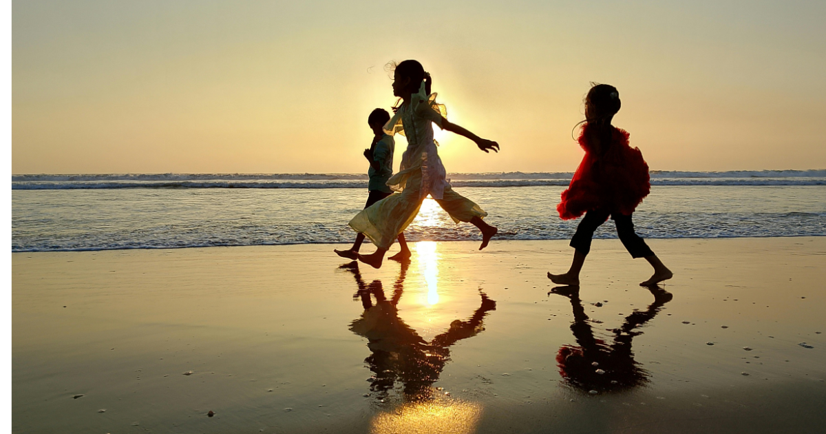 Children playing and running on the beach during sunset, with their reflections visible on the wet sand.