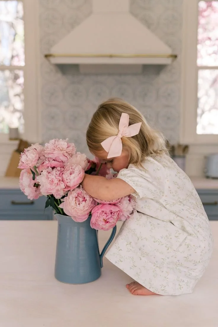 A young girl in a white dress with a pink bow in her hair is smelling pink peonies in a blue pitcher in a bright kitchen.