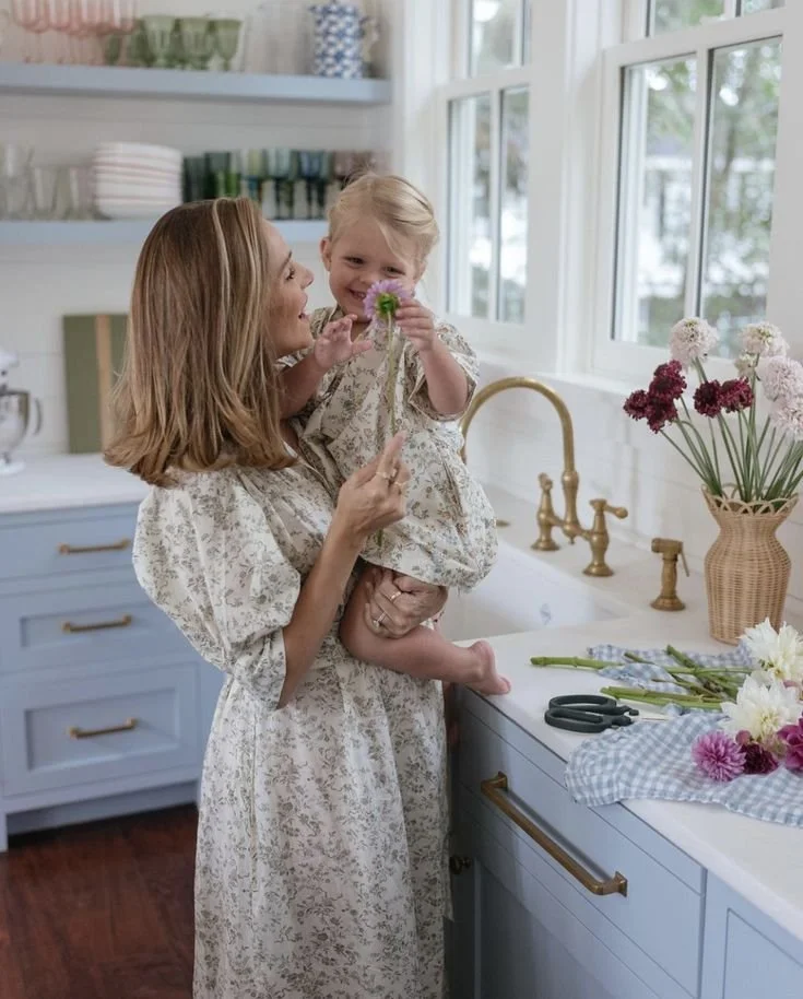 A woman holding a young girl in a kitchen, both smiling and holding flowers, with a vase of flowers on the counter nearby.