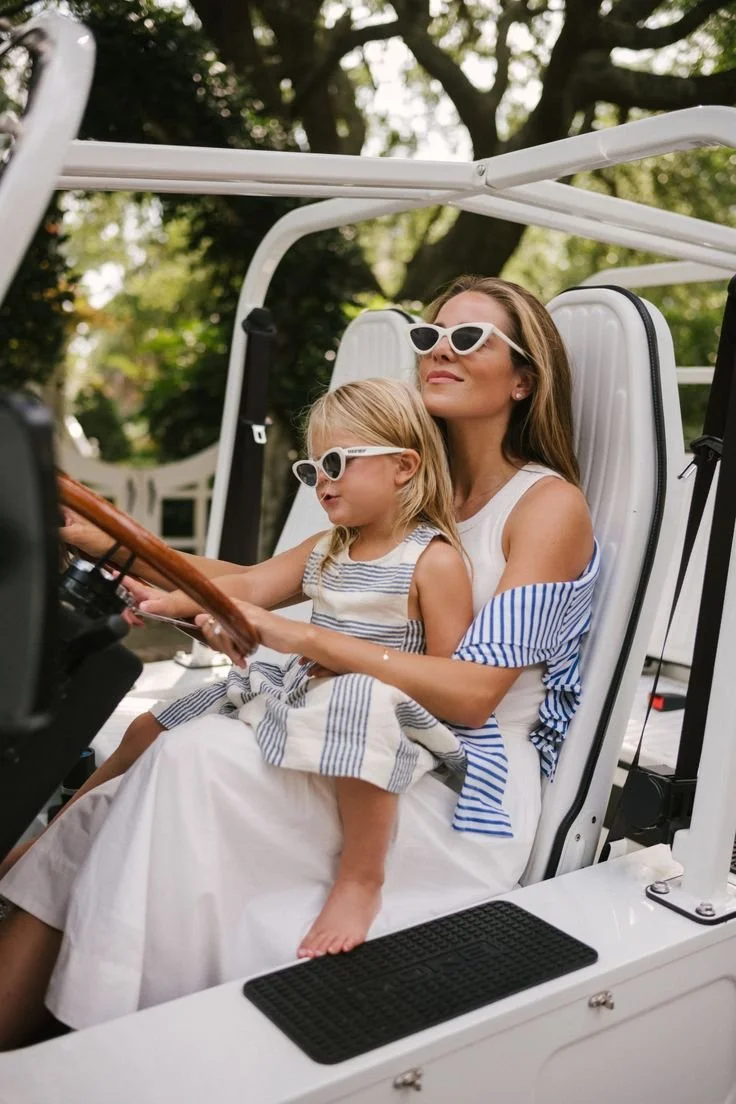 A woman and a young girl, both wearing white sunglasses, sitting in a golf cart outdoors with trees in the background. The woman is holding the steering wheel, and the girl is sitting on her lap, looking at something ahead.