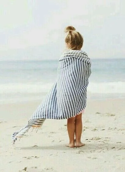 A young girl covered with a striped towel, standing on a beach facing the ocean.
