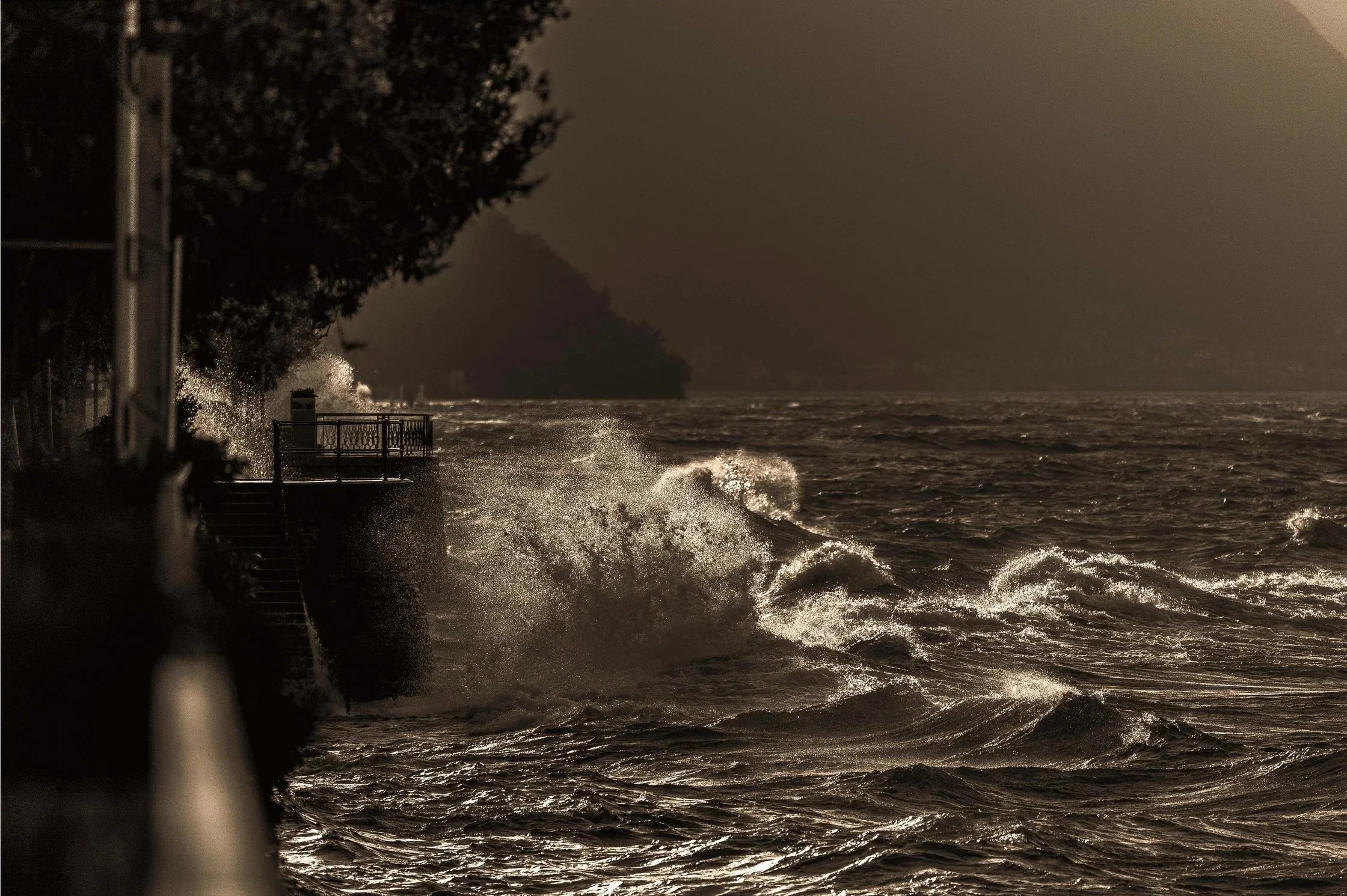 Nighttime scene of rough ocean waves crashing against a seaside walkway with a metal railing, silhouetted trees, and dark clouds in the background.