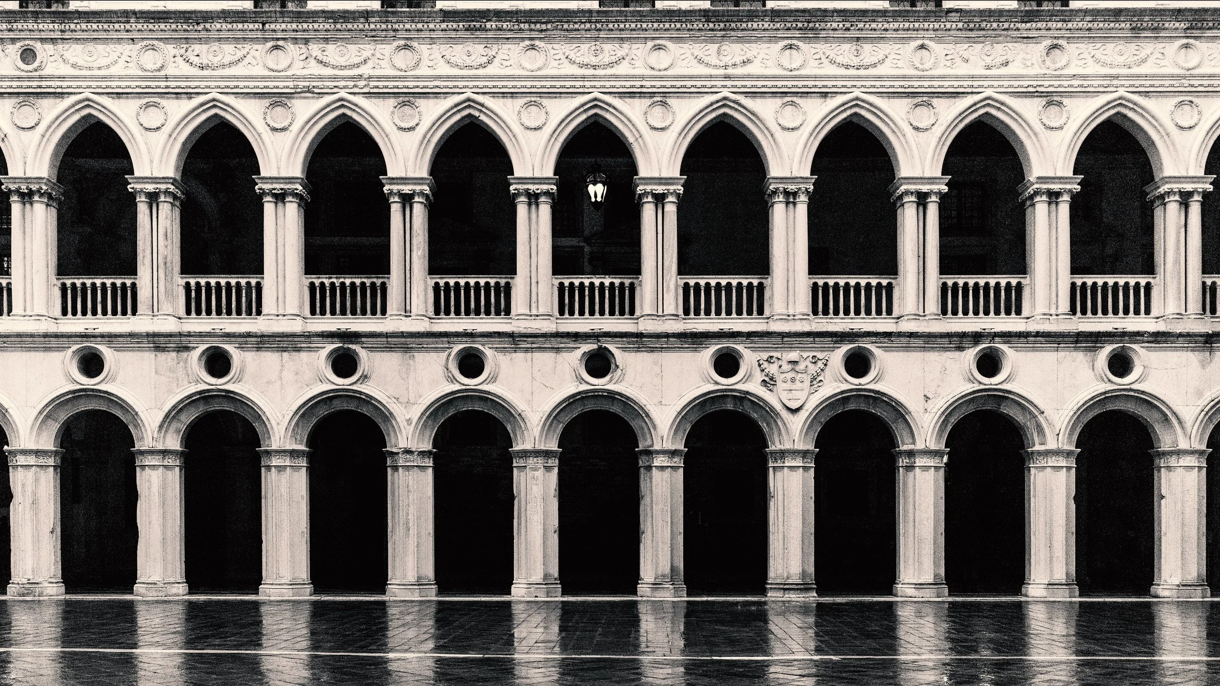 A black and white photograph of the façade of the Doge's Palace in Venice, Italy. The building features ornate architectural details, including a series of pointed arches on the upper and lower levels, supported by columns with decorative capitals.