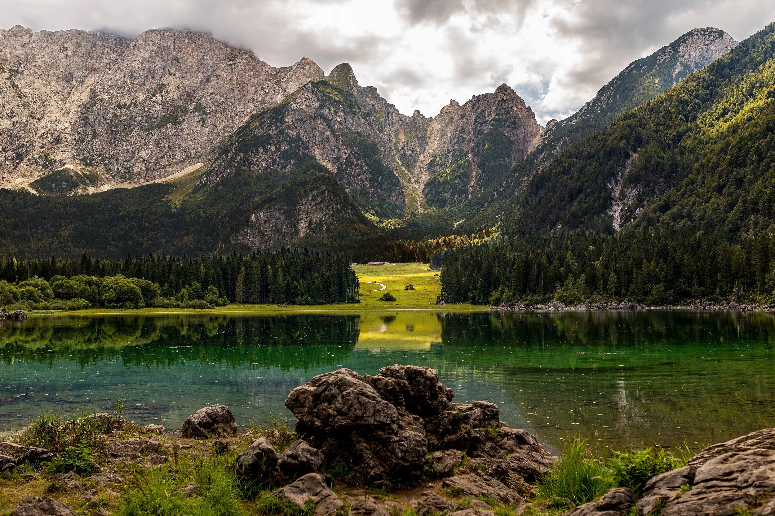 Mountains with forested slopes and a lake in the foreground, rocks along the shore, cloudy sky.