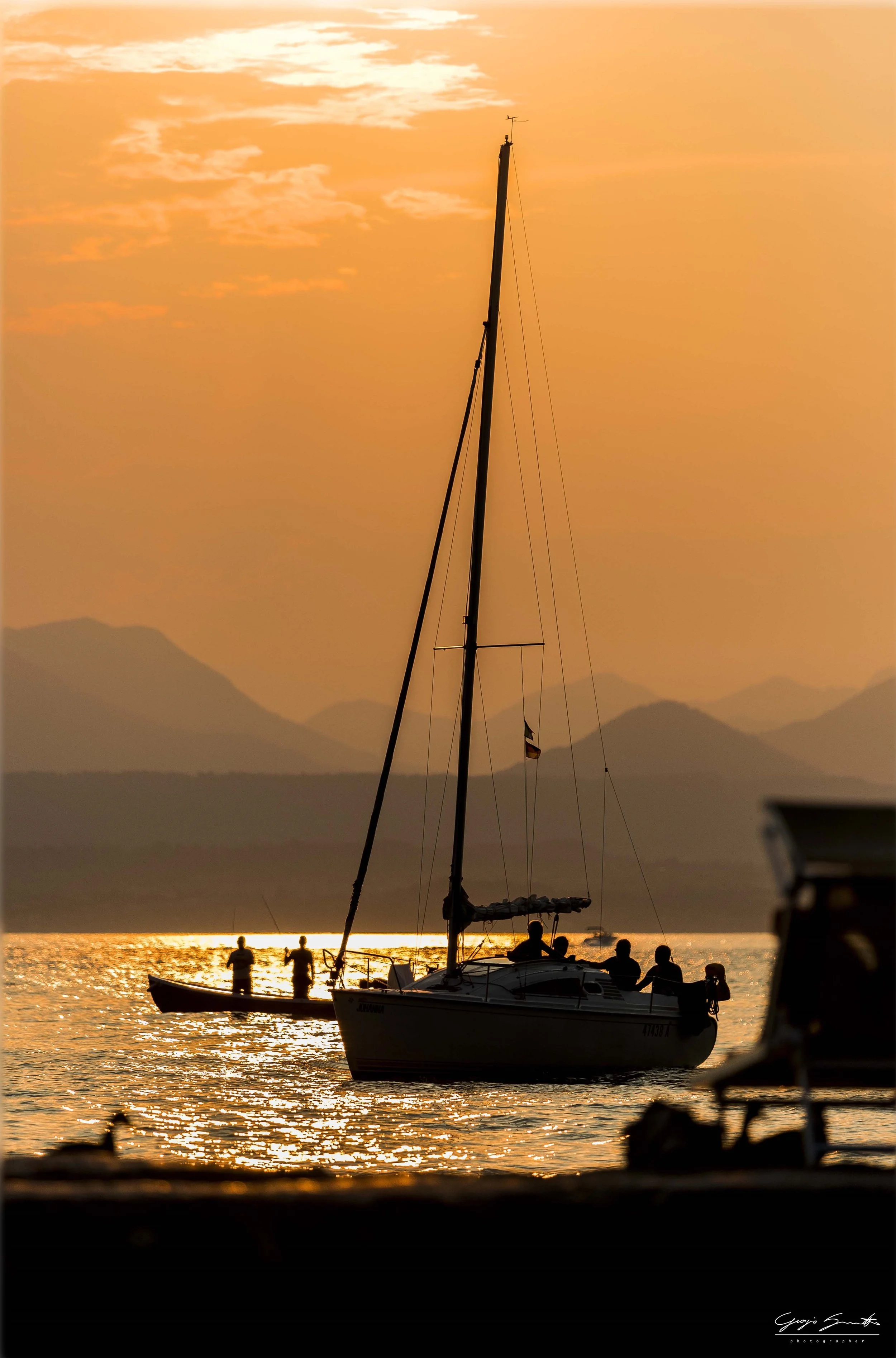 A sailboat with five people aboard on a body of water during sunset with a mountain range in the background.
