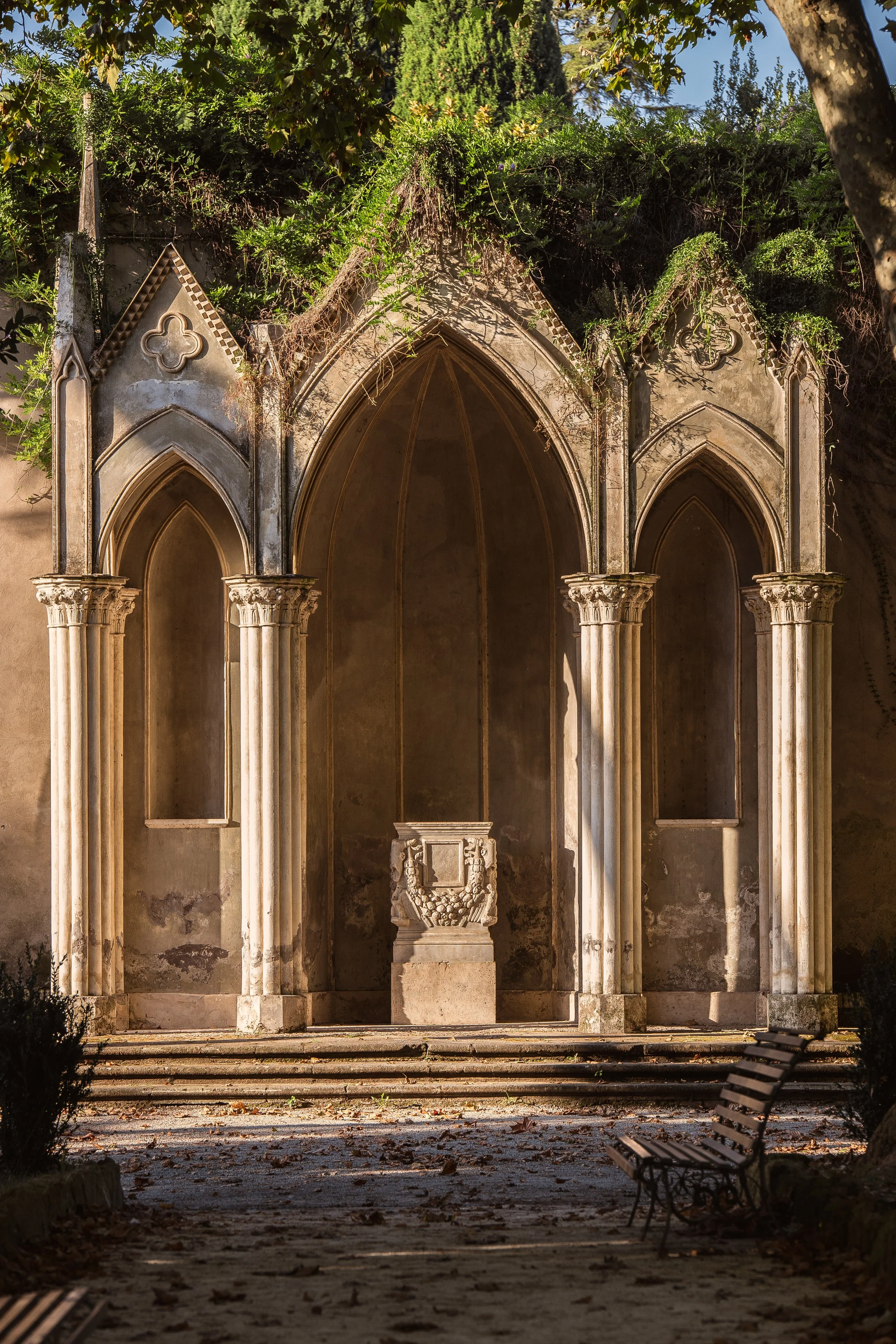 Fine art photograph of a sunlit Gothic cloister in Rome with pointed arches and marble columns, framed by overgrown greenery — Hermès Gray