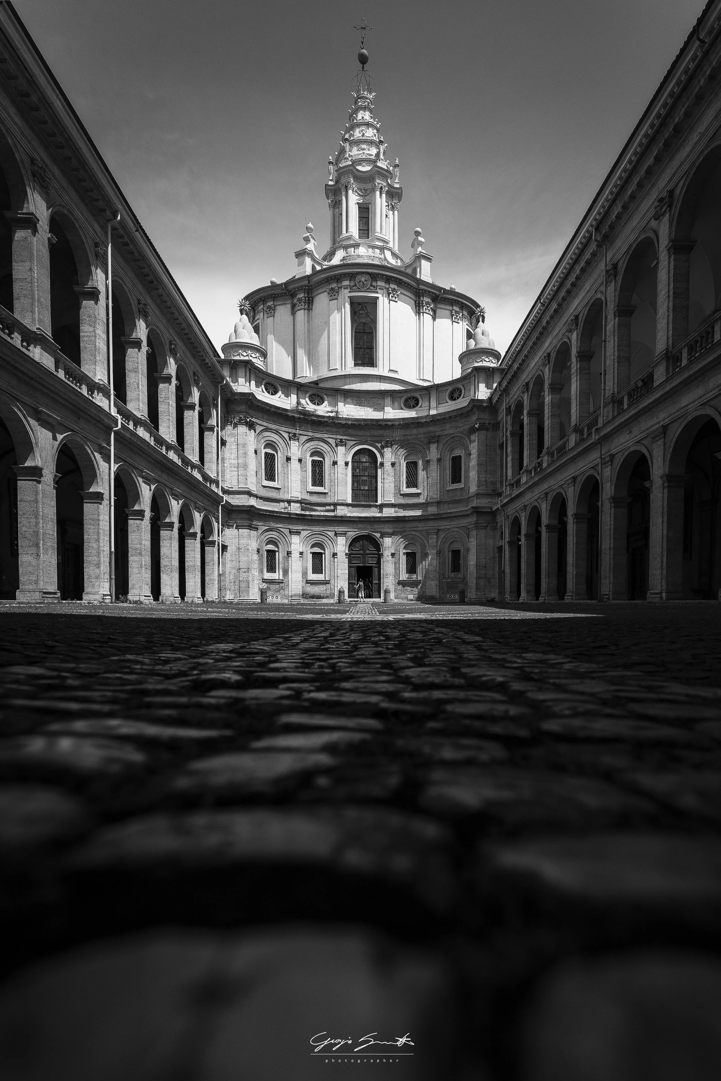Black and white photo of a baroque style church with a tall ornate steeple, viewed from the cobblestone courtyard below.