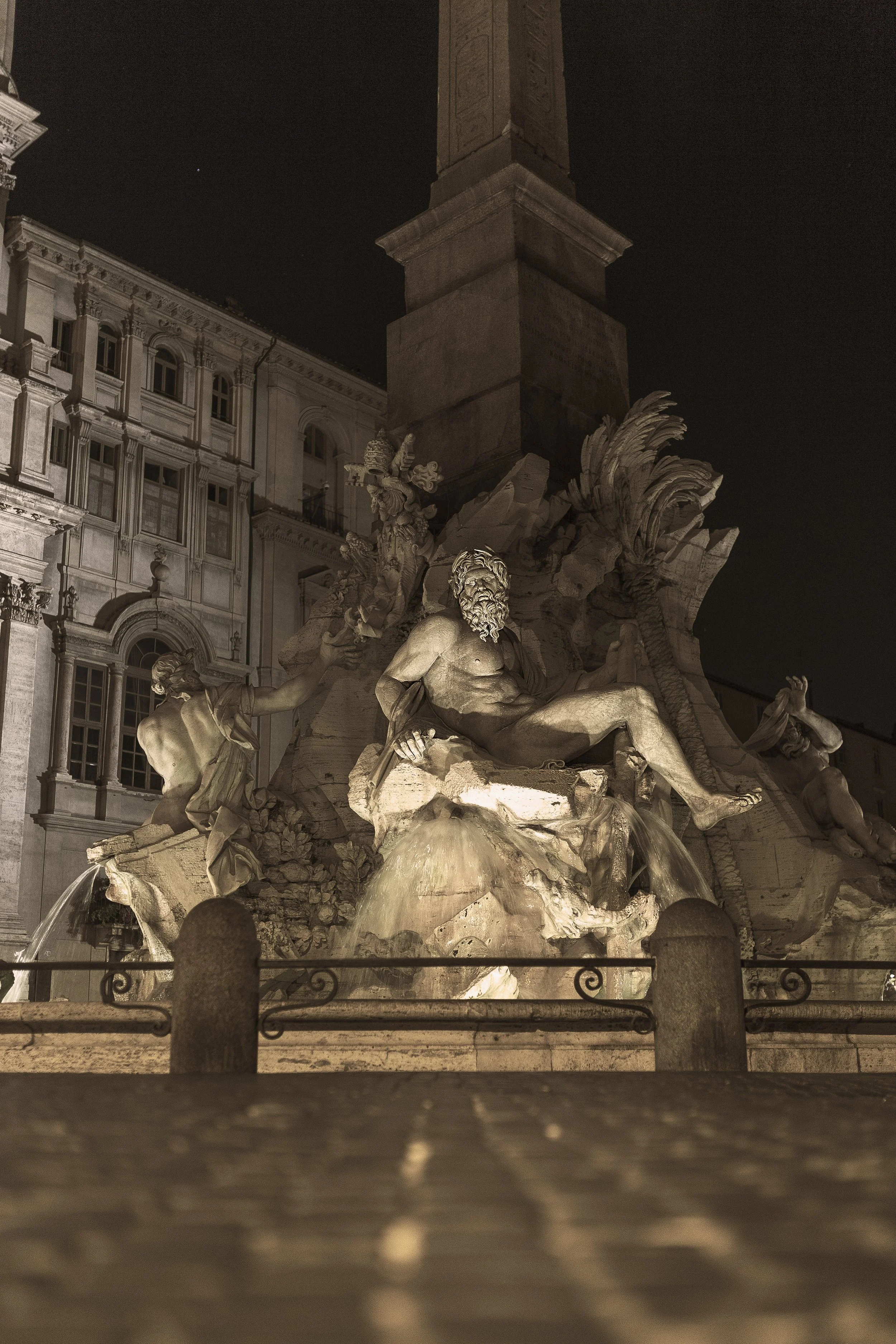 Night view of the Fountain of the Four Rivers in Rome, Italy, showing statues of rivers and the obelisk in the center, illuminated against a dark sky with buildings in the background.