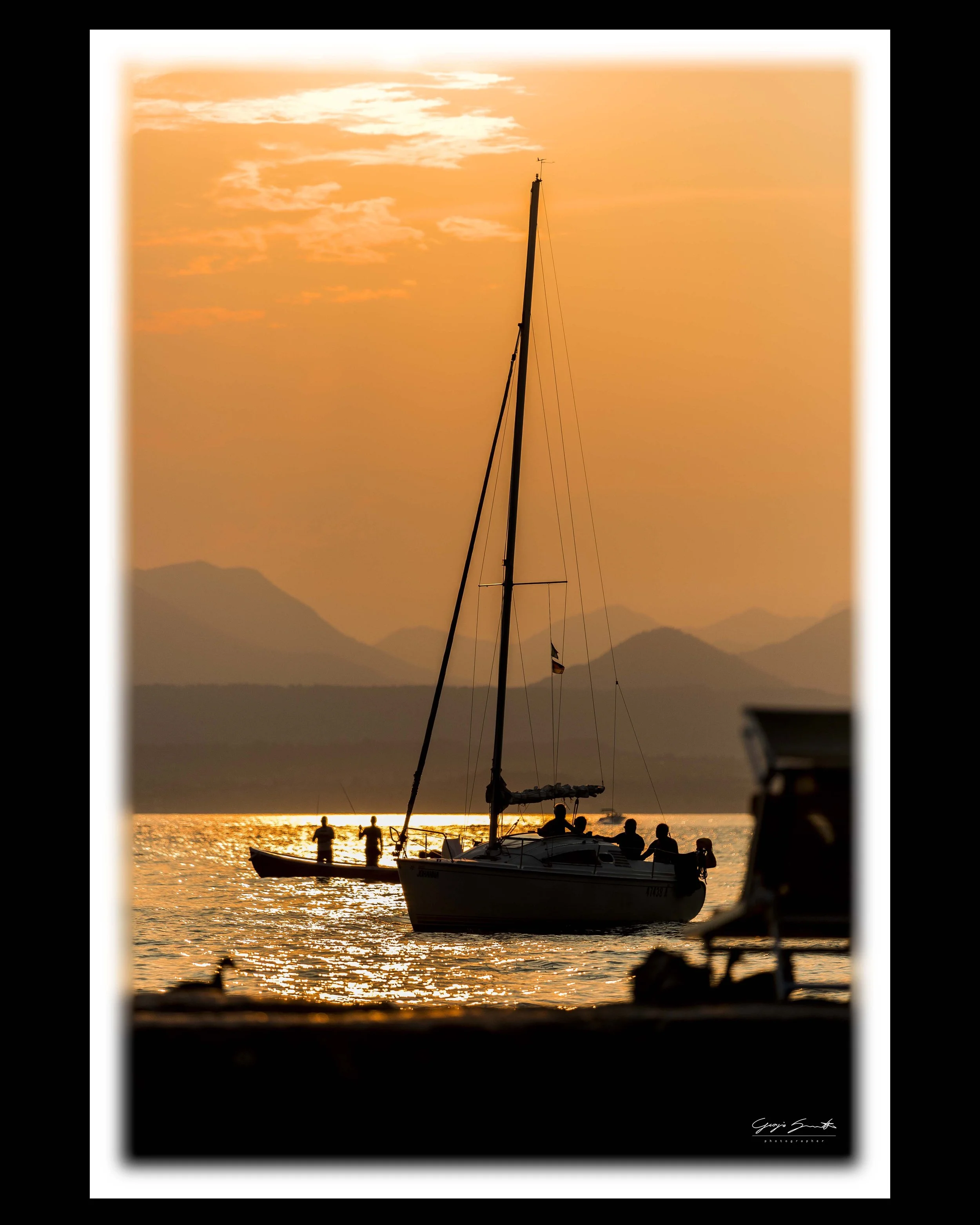 Sailboat silhouette at golden hour, figures on deck reflected in calm water, mountains in background. Fine art limited edition print, Mediterranean sunset photography.