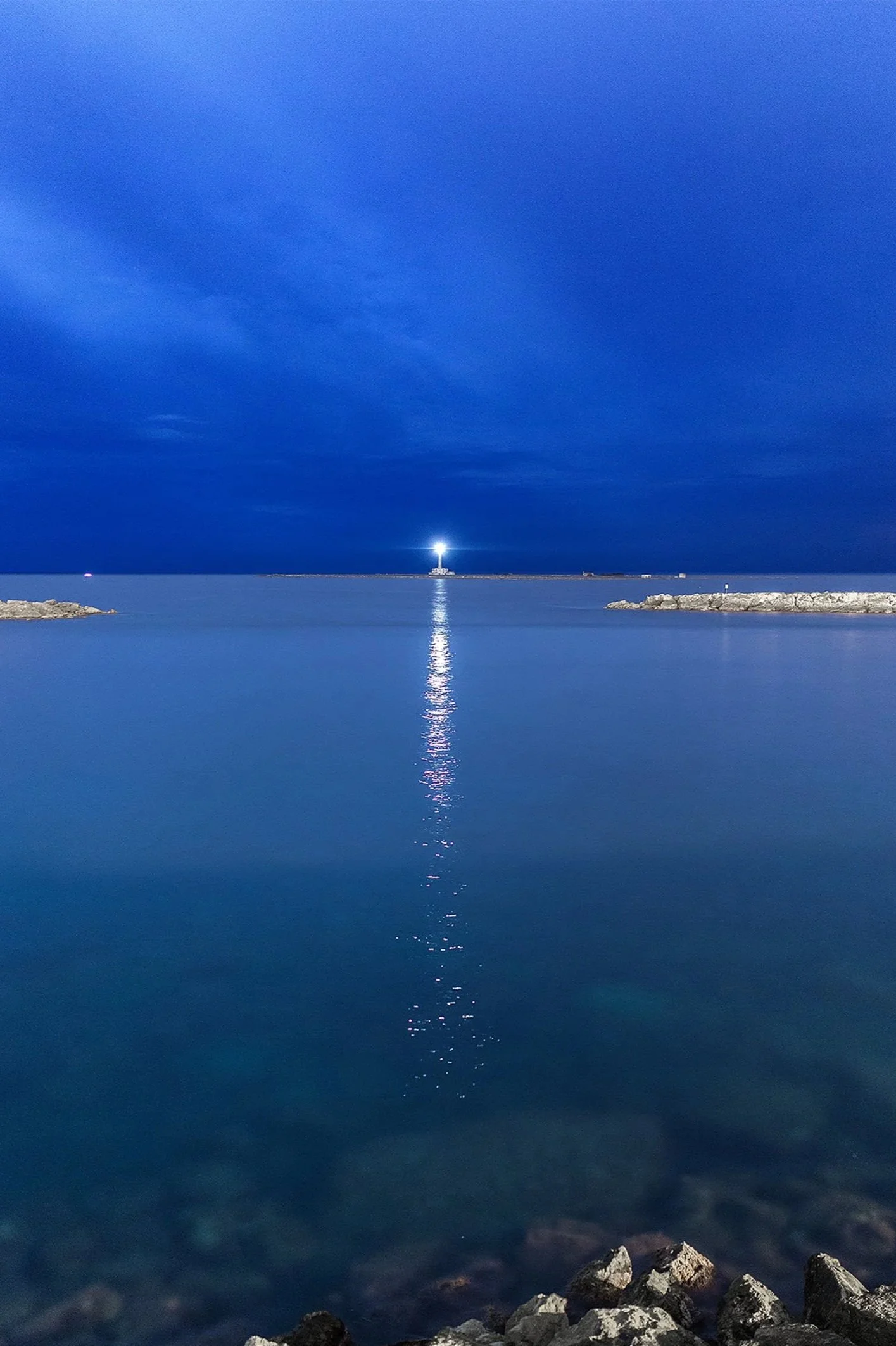 Nighttime view of a lighthouse on a small island, casting a bright light across the water, with a reflection visible on the calm ocean surface, and rocky shoreline in the foreground.