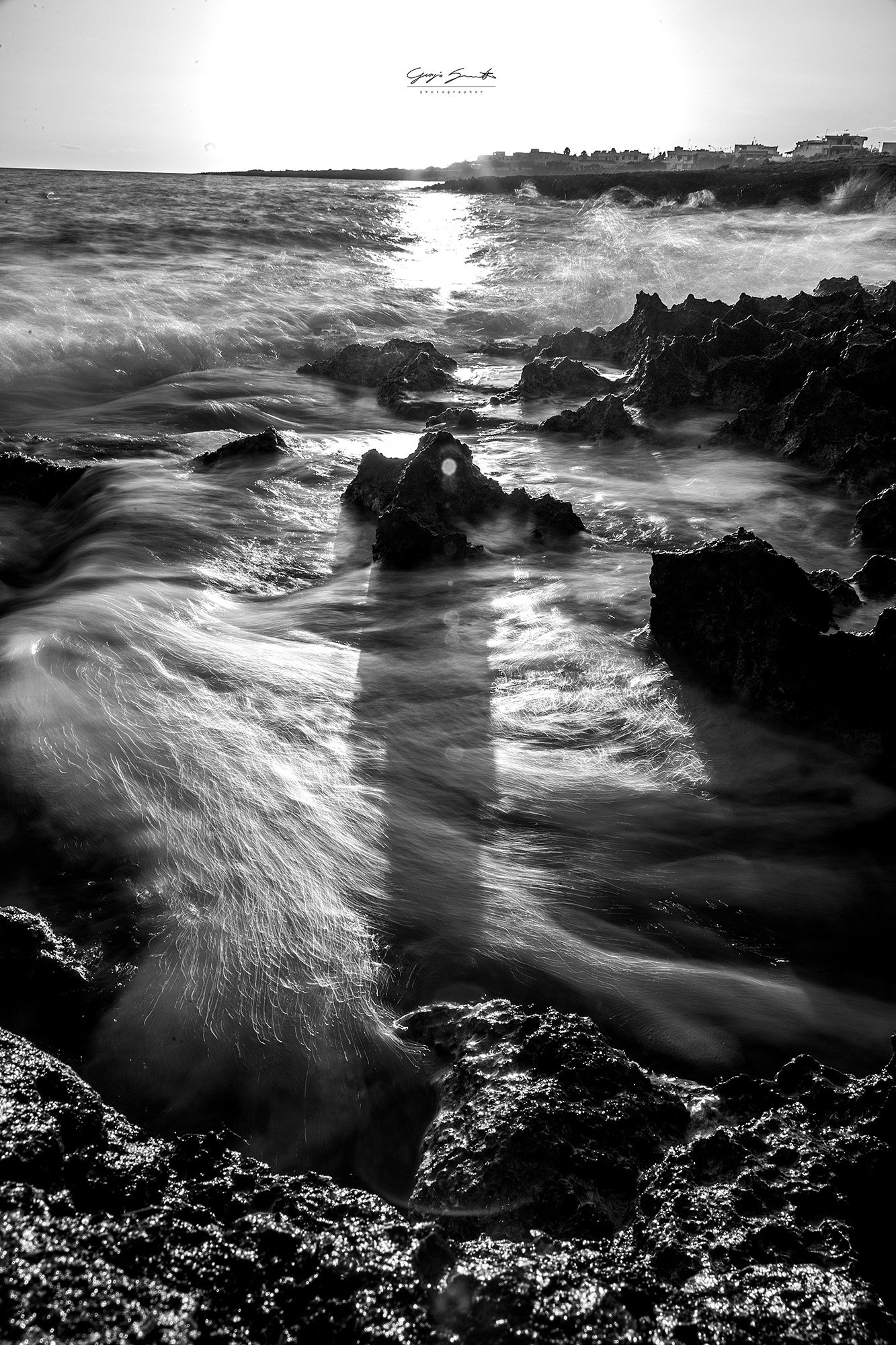 Black and white photograph of ocean waves crashing against rocks on the shoreline, with buildings visible in the background.