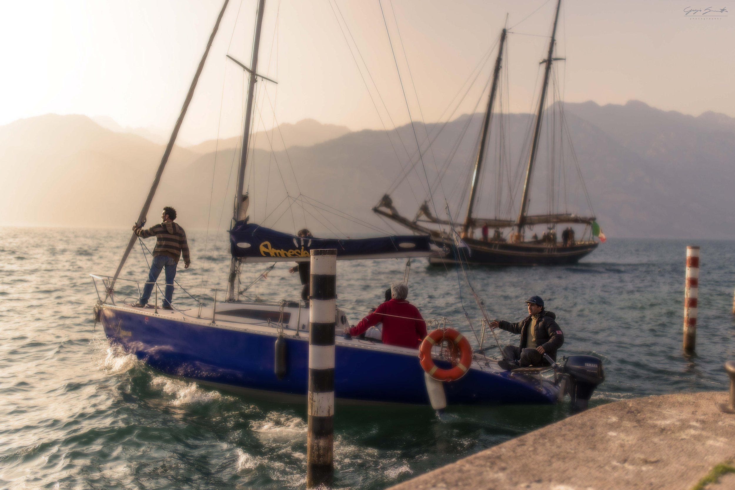 Sailors maneuvering sailboats at a misty harbor at golden hour, Lake Garda, Italy. Documentary fine art photography, limited edition print on Hahnemühle paper.